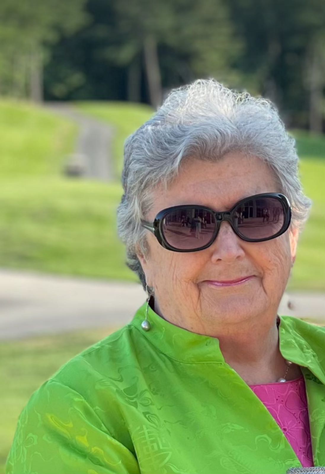 Brenda O’Connor, my mother, wearing a green jacket over a pink blouse. She has sunglasses on, and is smiling somewhat pensively at the camera.