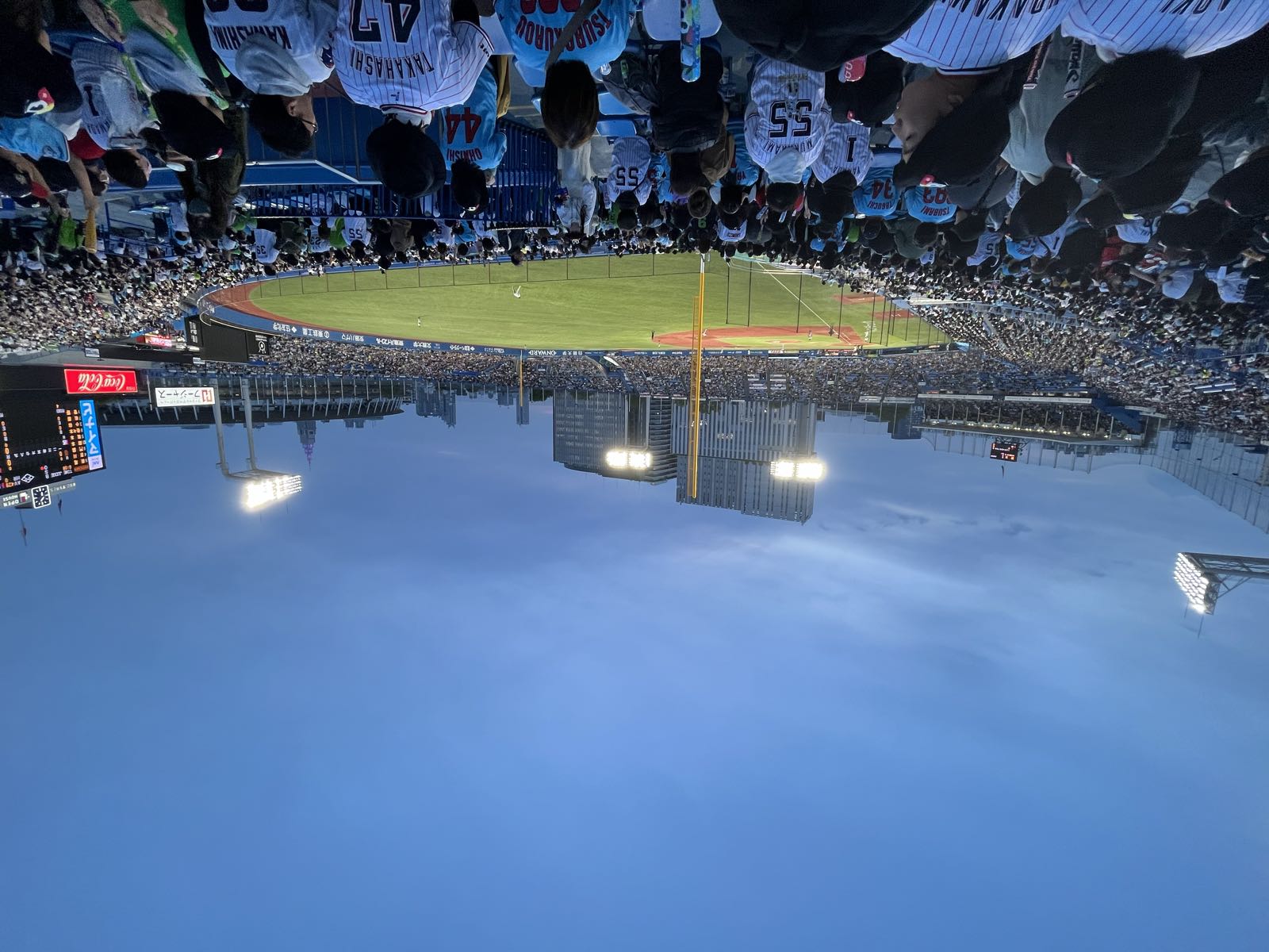 Baseball at night in Tokyo.
A packed Meiji Jingu Stadium is lit up by large banks of lights.
The photo takes in most of the stadium from the right field stands, just foul of the pole.
Docomo Tower looms over the stadium in left field.
Many of the people in the stands in front of the photographer are wearing Yakult Swallows jerseys.