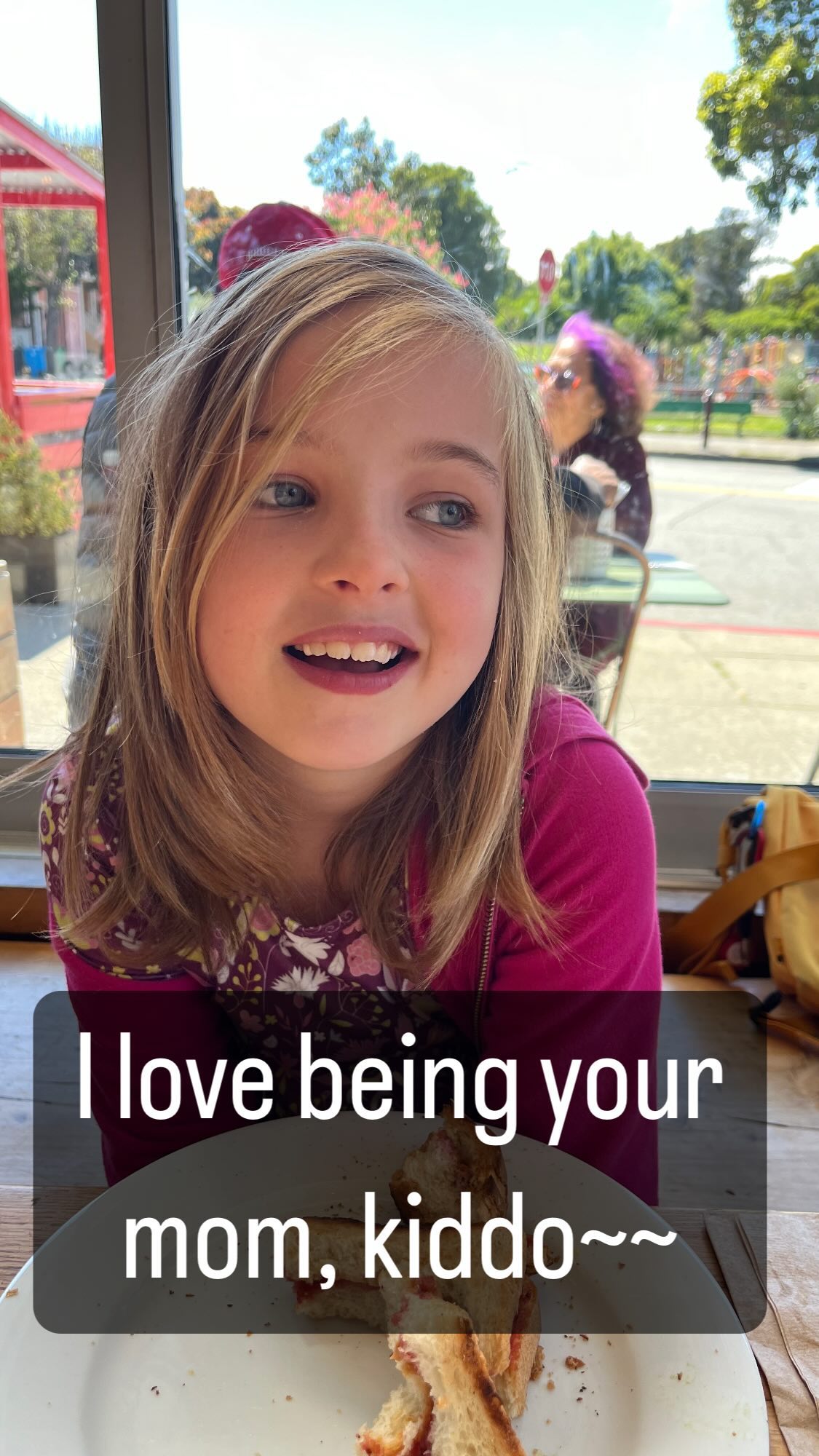 A smiling girl sits at and leans on a table.
In front of her is a plate with a mostly-eaten sandwich on it.
She’s wearing a floral dress with a pink cardigan over it.
Her straight, dirty blonde hair extends past her shoulders.
In front of her is text that reads “I love being your mom, kiddo~~”.