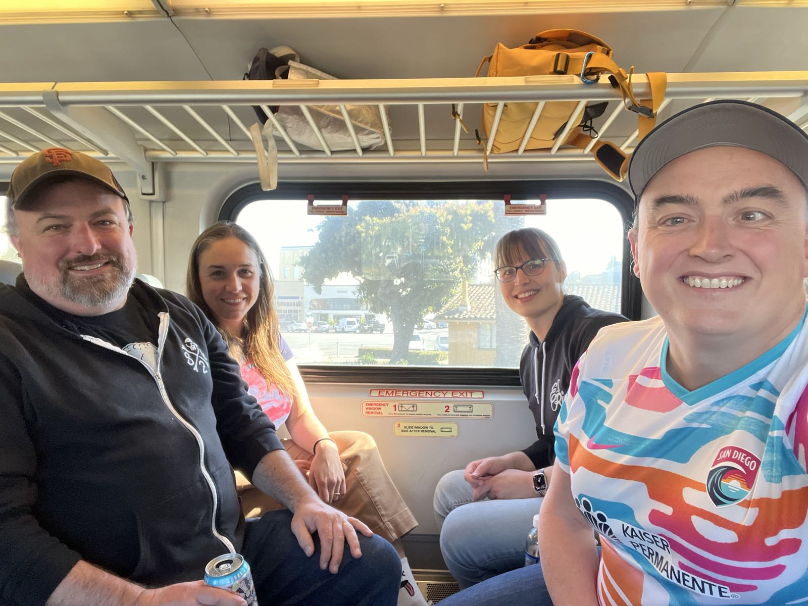 Four adults sit facing each other on a train. Everyone is smiling. The man on the left is in a San Francisco Giants hat and a black hoodie. Next to him, a woman with long hair sits next to the window. Across from her, a woman with glasses is also wearing a black hoodie. And to her right, a smiling woman is wearing a San Diego Wave jersey.