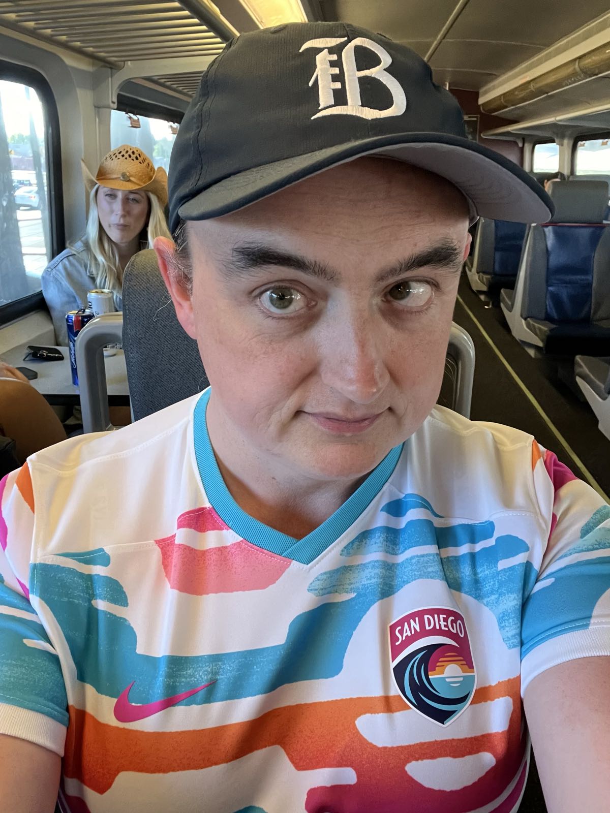 A selfie of a woman. She’s wearing a navy blue Bay FC hat and a white San Diego Wave jersey. She has severe strabismus.
