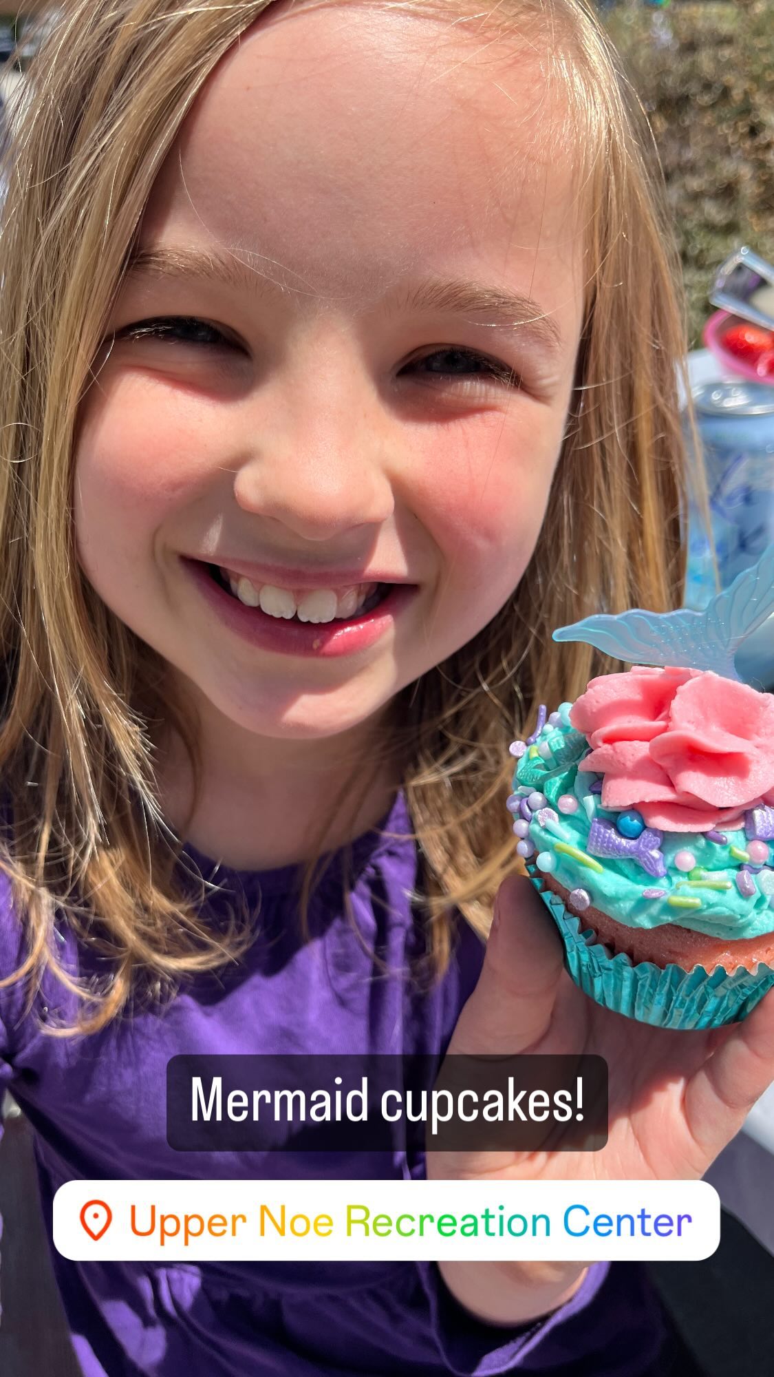 A smiling child holds a pastel-frosted cupcake up to the camera.
She has dirty blonde hair.
Two sets of text are printed on top of the image.
The first reads “Mermaid cupcakes!”, and the second, “📌 Upper Noe Recreation Center”.