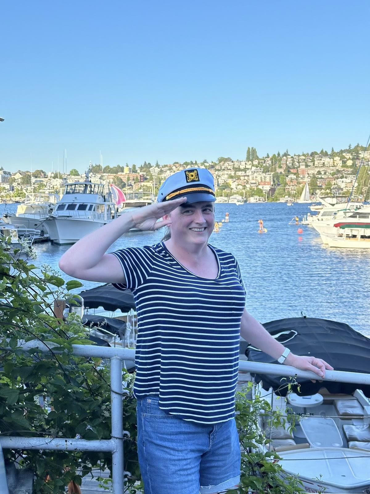 A goofy-looking woman in a blue-and-white striped T-shirt, denim shorts, and a captain’s hat is smiling and saluting the cameraperson.