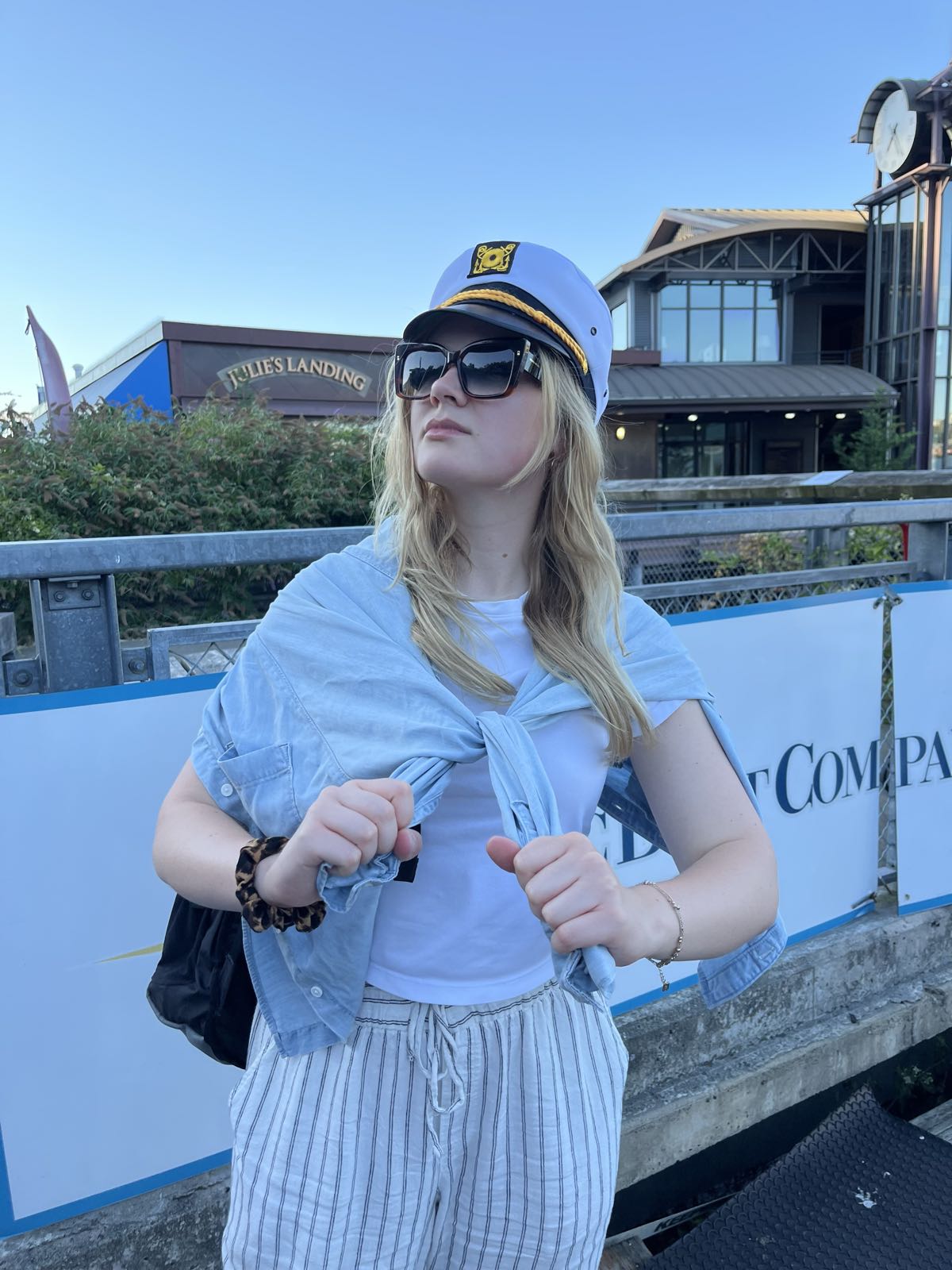 A woman in a T-shirt has a button-up shirt tied around her neck.
She’s wearing sunglasses and is looking off to the left.
She’s wearing a captain’s hat.