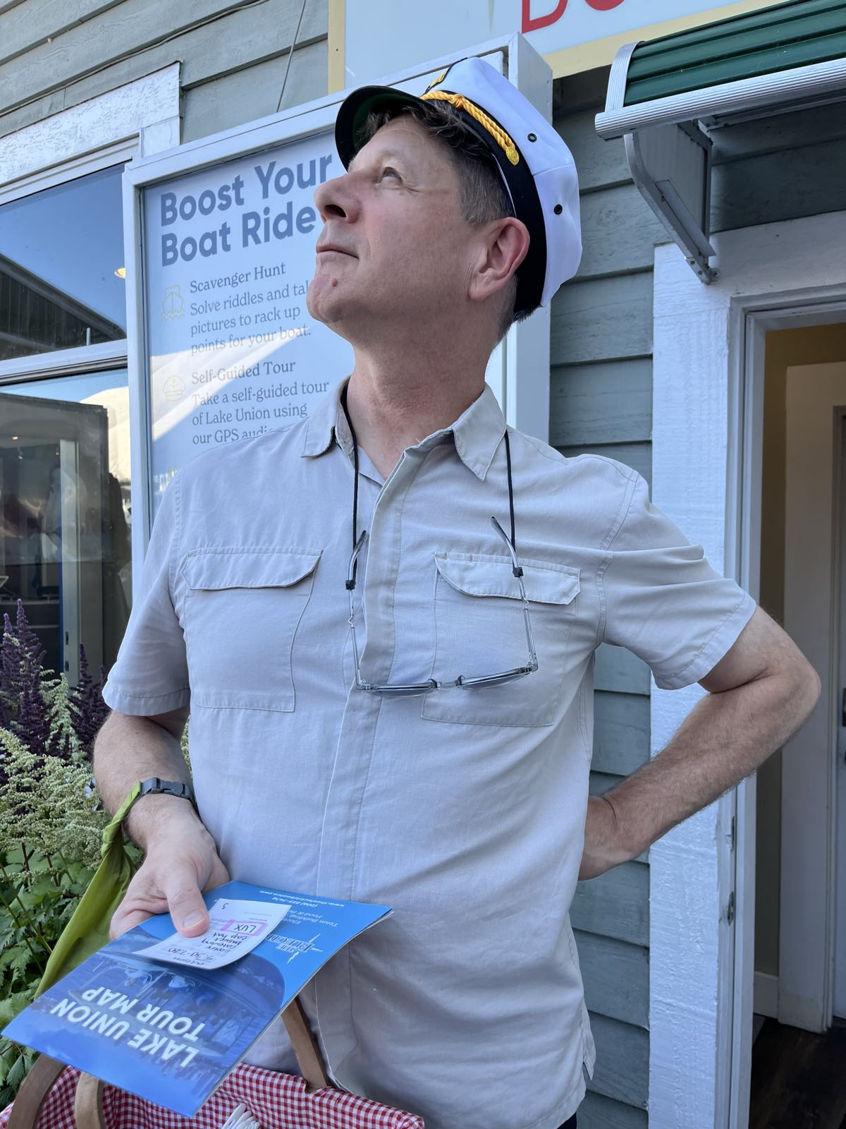 A man strikes a haughty pose while wearing a captain’s hat. In his right hand he holds some papers. Around his neck hang a pair of glasses.