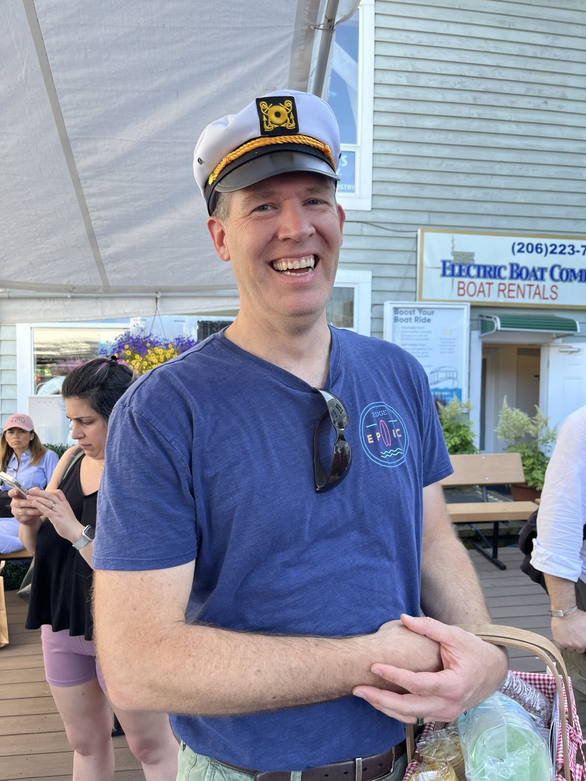 A man in a blue T-shirt and a captain’s hat smiles at the camera. His hands are clasped in front of him. A pair of sunglasses hang from his collar.