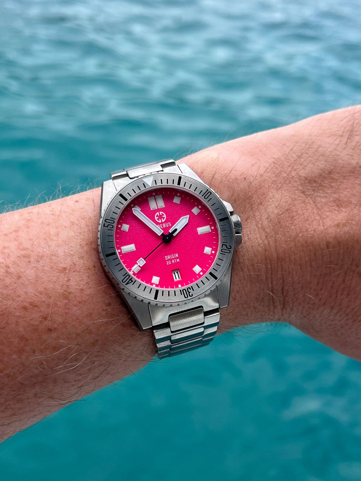 A person is wearing a steel dive watch with a vibrant pink dial on their left wrist.
They’re holding up their arm in front of a pretty body of water on a sunny day.