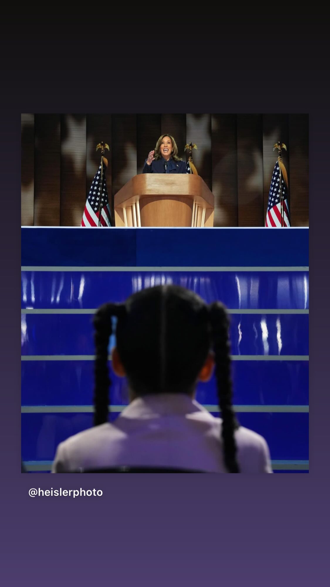 A photo of Kamala Harris giving her acceptance speech at the DNC last week. In the foreground, a young girl watches her.