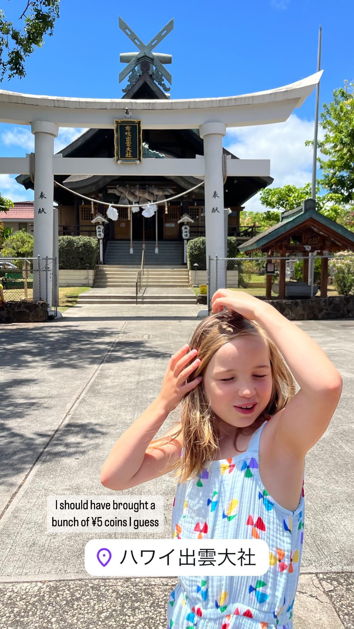 Erin’s hands are in her hair and she’s making a strange face while standing in front of a Shinto shrine with a large, white torii gate.