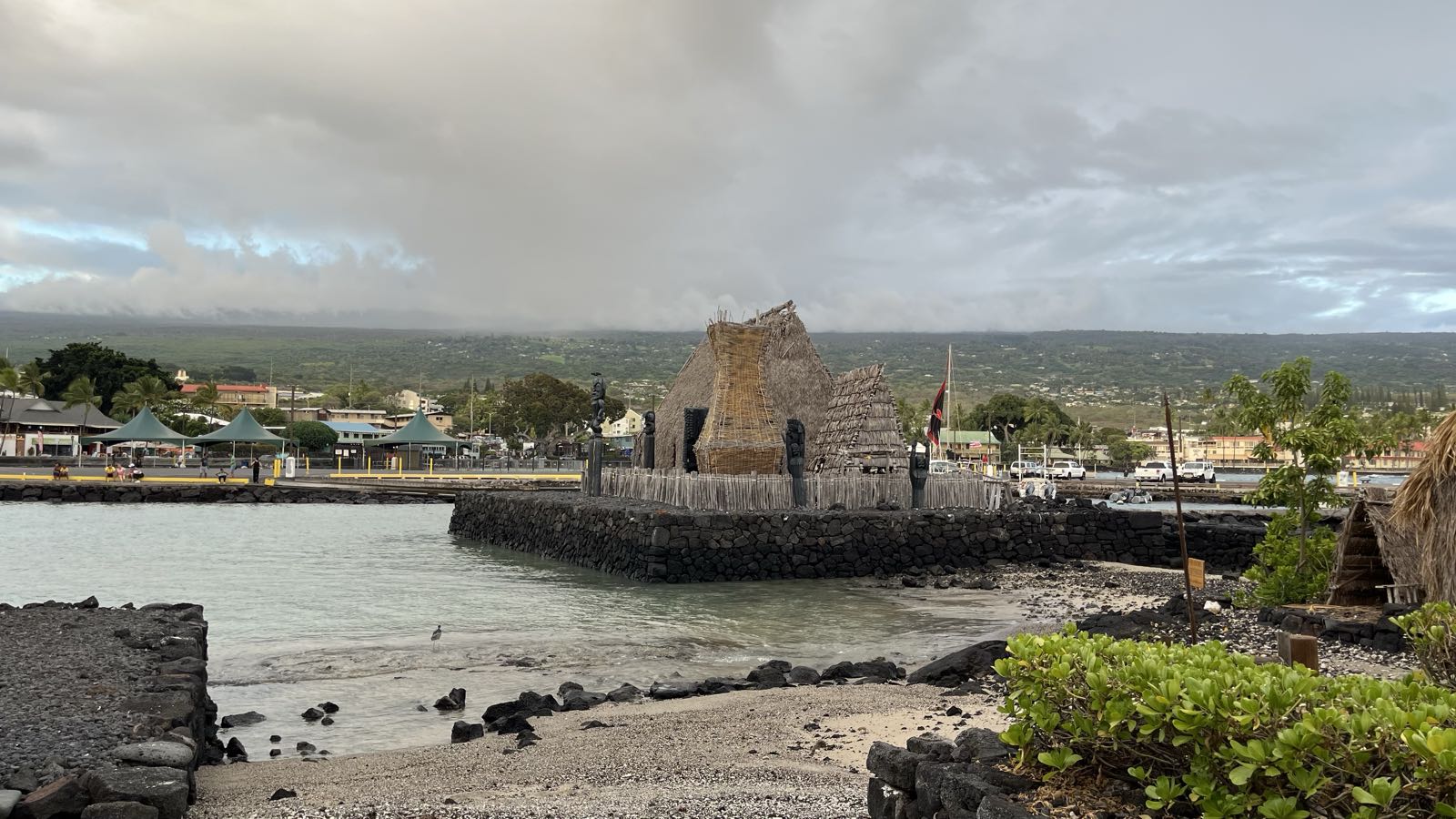 An ancient-looking straw structure stands on dark, igneous rock in the middle of an idyllic harbor.