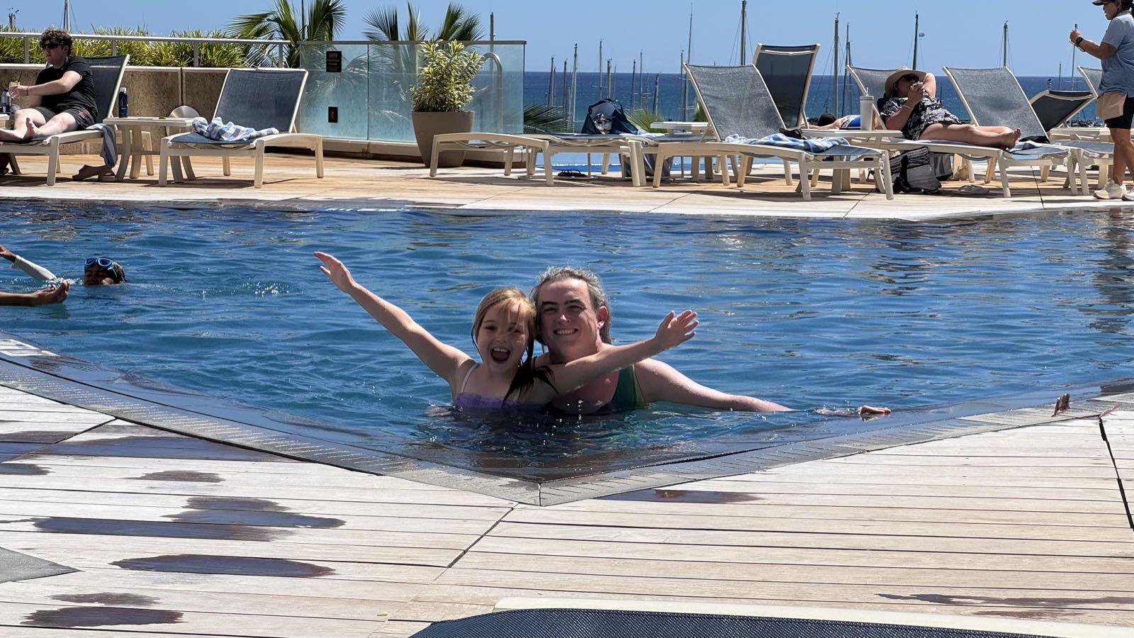 A smiling adult and child stand in a swimming pool. The child’s arms are outstretched in joy.