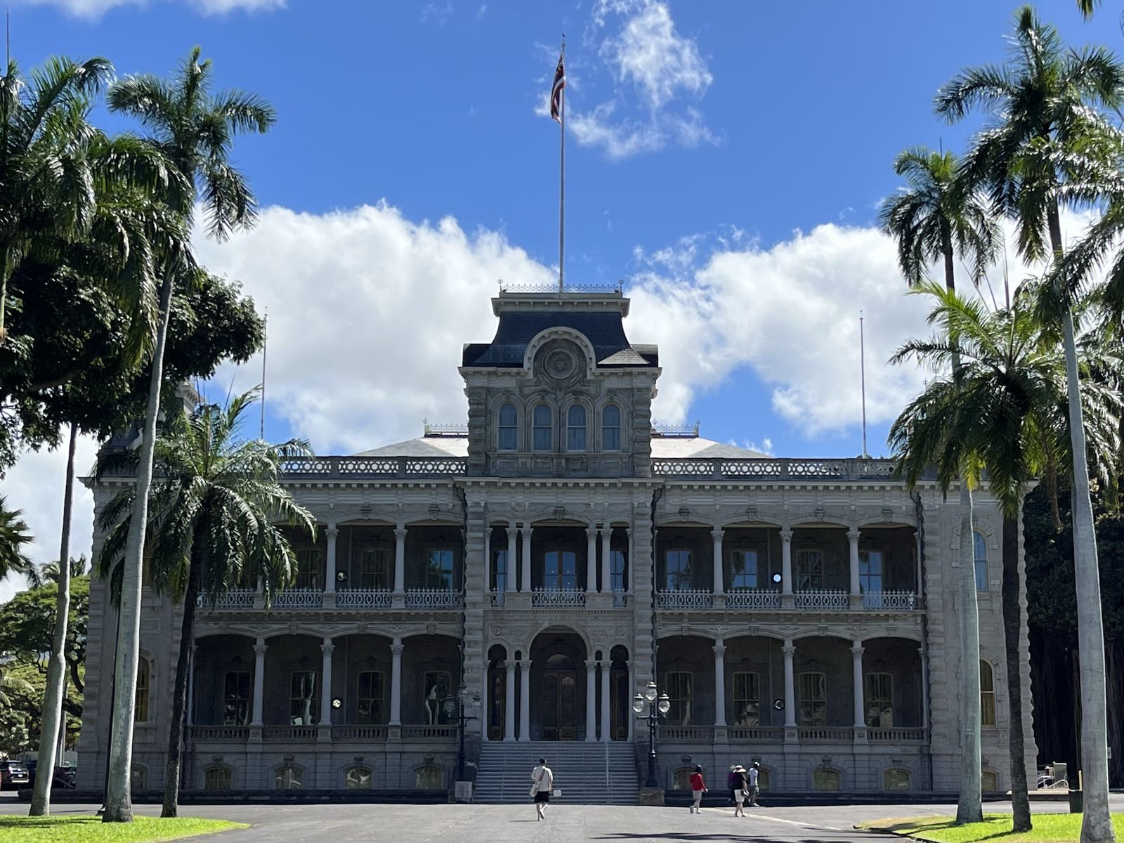 The facade of ʻIolani Palace.
The palace is two stories, with a central tower of three.
There are palm trees to either side of it.
The American flag is flying from its flag pole.
