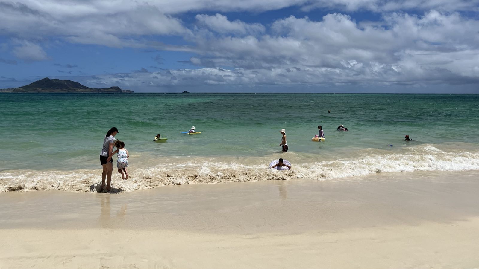 Eryn helps EJ jump over waves at Kailua Beach on the windward side of Oahu. There are a number of other people in the water, with boogie boards and inner tubes.