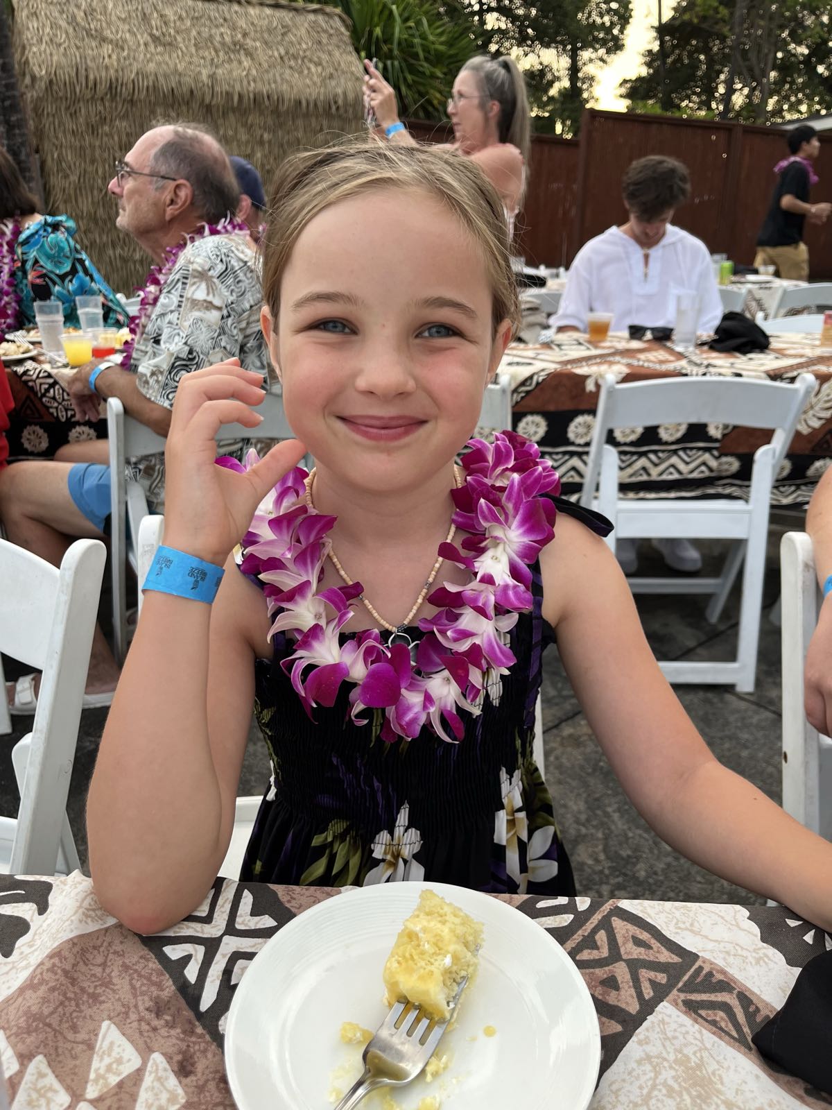 Erin is sitting at a table.
She’s in a dark, floral dress, and has a purple and white lei draped around her neck.
There’s a half-eaten piece of white cake on her plate.