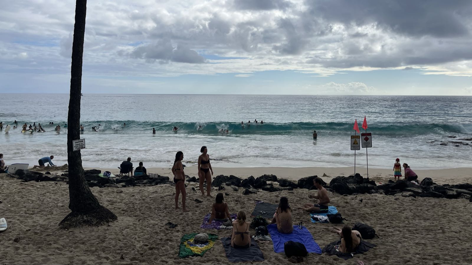 A wave is about to break at a beach.
A number of people are attempting to ride the wave with boogie boards.
Other people sit or stand on the beach and watch.
The sun is obscured by clouds.
