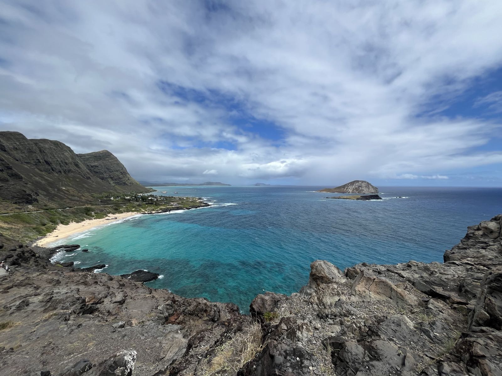 The Pacific Ocean as seen from Makapuʻu Lookout.
The sky is cloudy. There’s a beach along the left side of the shot. There is another island visible out in the water.