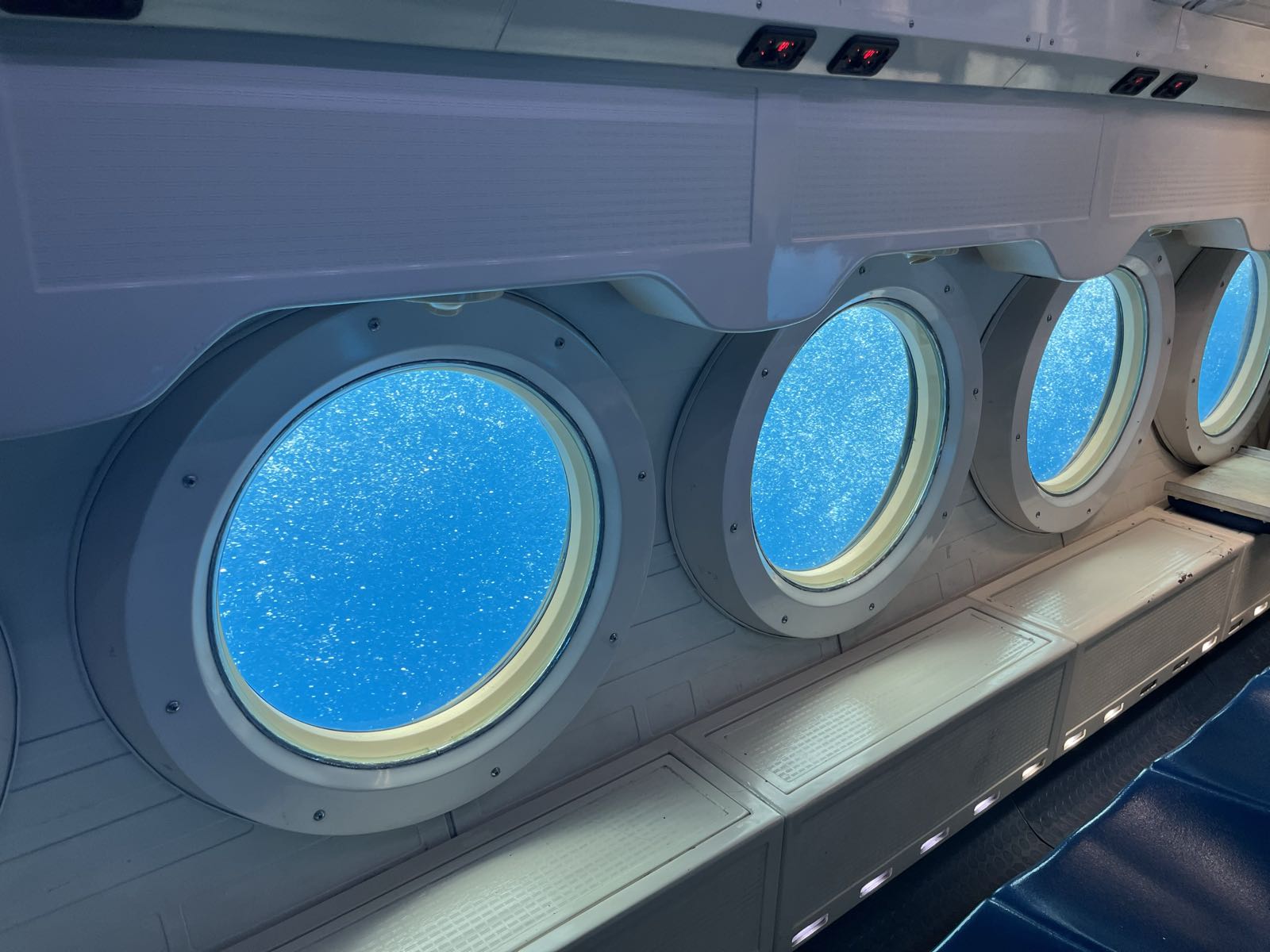 A row of four portholes reveal bubbly ocean water beyond the interior of a submarine. Blue seating is aligned with the portholes.