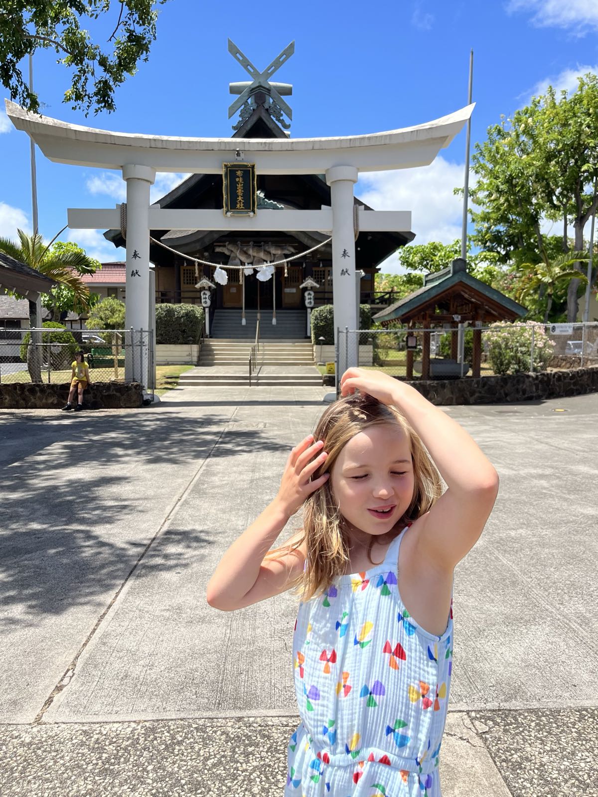 Erin’s hands are in her hair and she’s making a strange face while standing in front of a Shinto shrine with a large, white torii gate.
