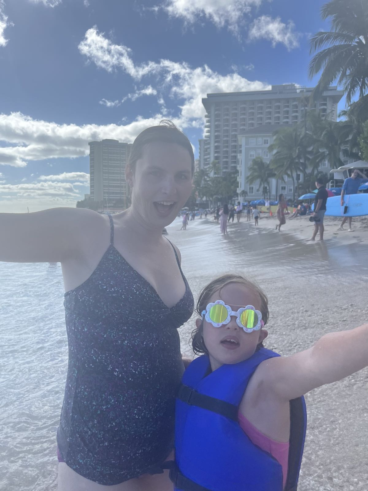 Eryn and EJ at Waikiki Beach.
Both have their arms outstretched and are making silly faces.
Eryn is in a dark, speckled, one piece bathing suit.
EJ has flower-shaped goggles on, as well as a blue life jacket.
Underneath the life jacket, her bathing suit is pink.