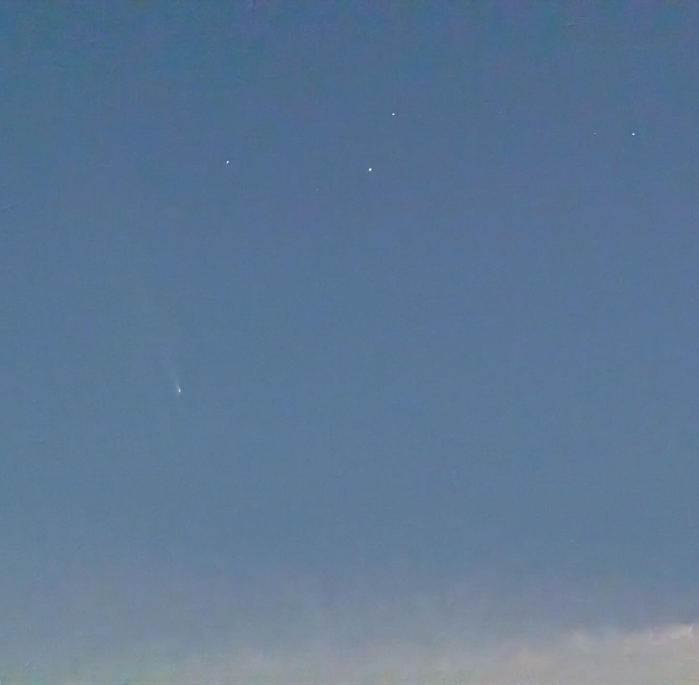 A blue sky, after sunset but before darkness has completely fallen. A half a dozen stars are visible. In the center left, a comet is visible.