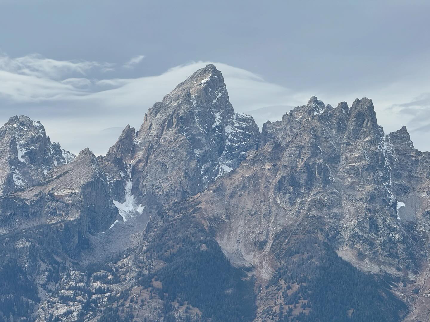 The peaks of the Grand Tetons are gray, with some snow here and there. The sky is also gray, and somewhat cloudy.