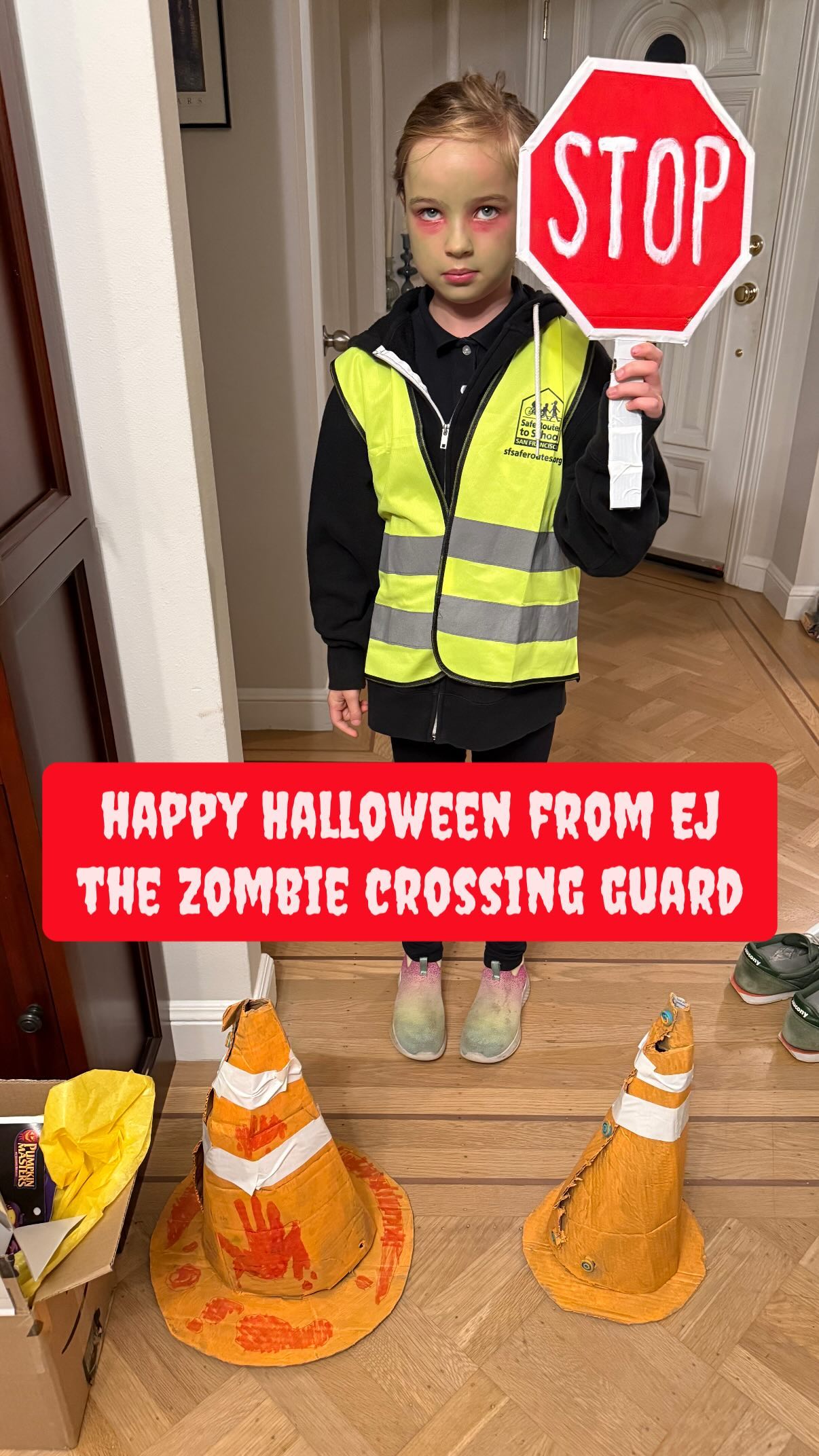 A child with sickly-looking green and red face paint wears a yellow-green reflective safety vest and is holding up a red, octagonal stop sign. In front of them are two orange safety cones, one of which appears to have bloody markings on it, including a hand print.