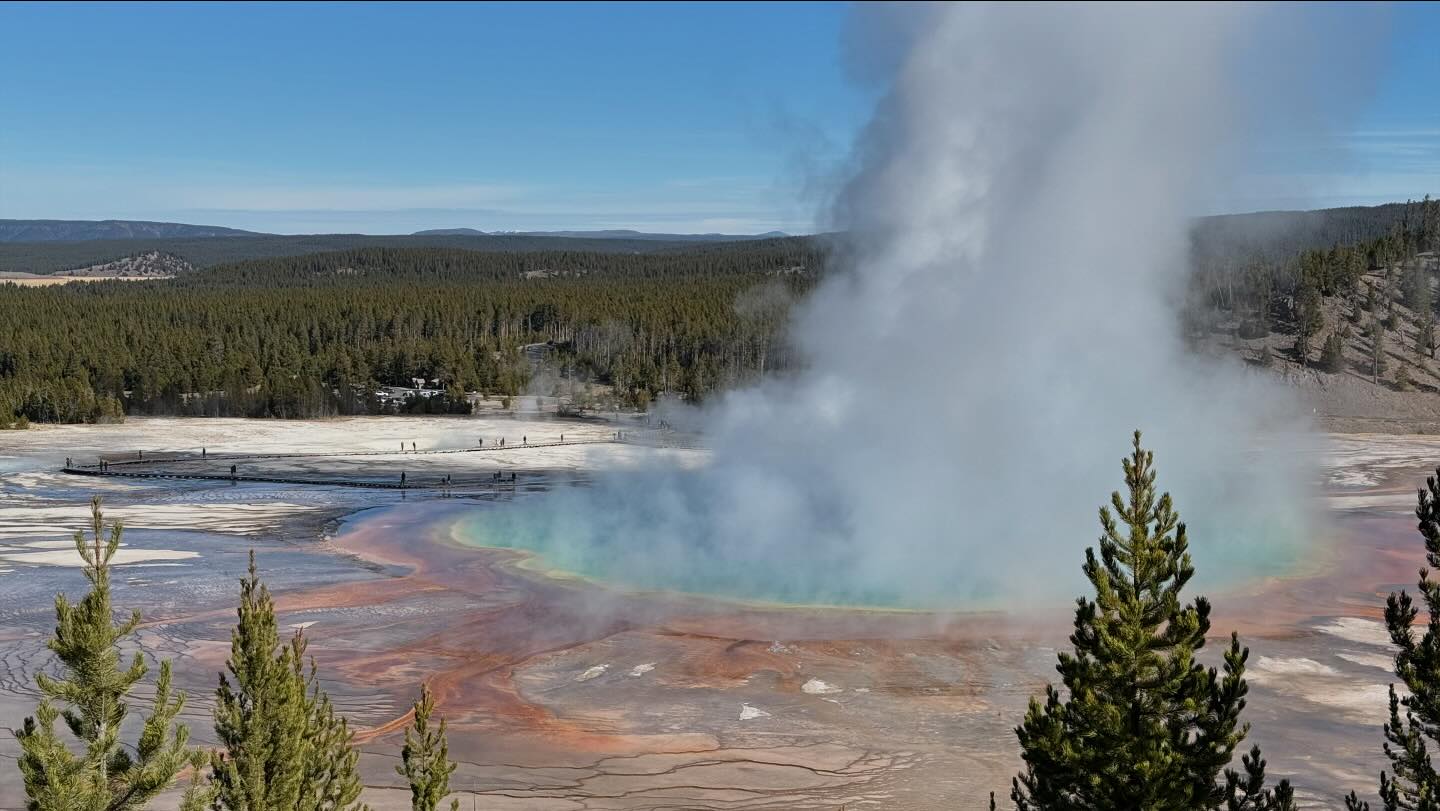 A large, circular hot spring is steaming. The water is a vibrant blue-green, and the ground around the spring is a colorful explosion of reds, oranges, and yellows.