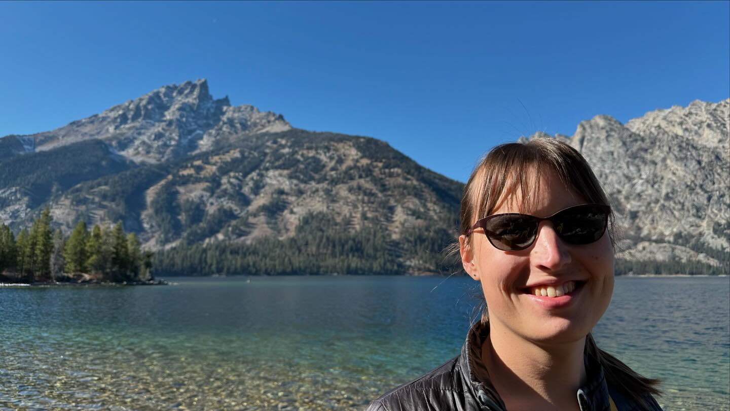 A smiling woman is standing in front of a lake. Beyond the far shore of the lake are two mountains. The woman is wearing sunglasses and a black puffy jacket.