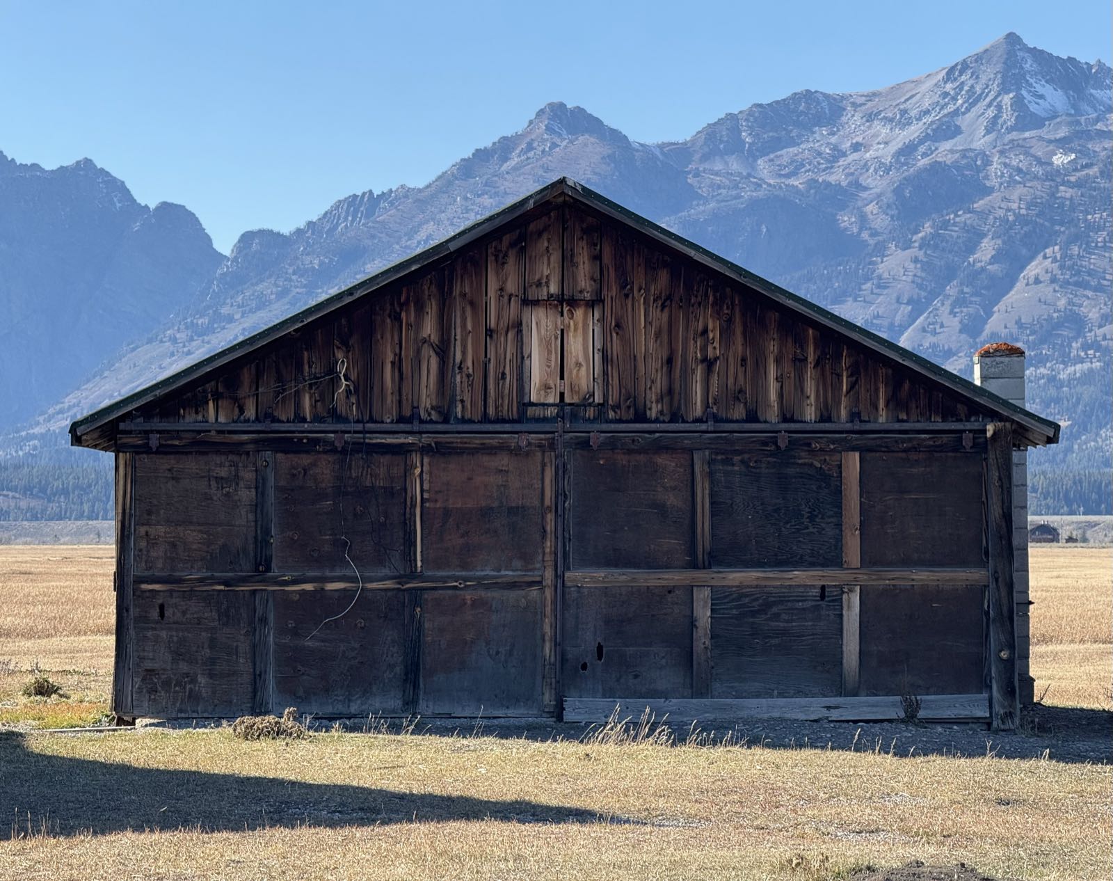 A barn stands in a field below mountains. The slope of the barn roof is lined up with the rise of the mountain range behind it. The field is covered with yellow grasses. The barn appears abandoned.