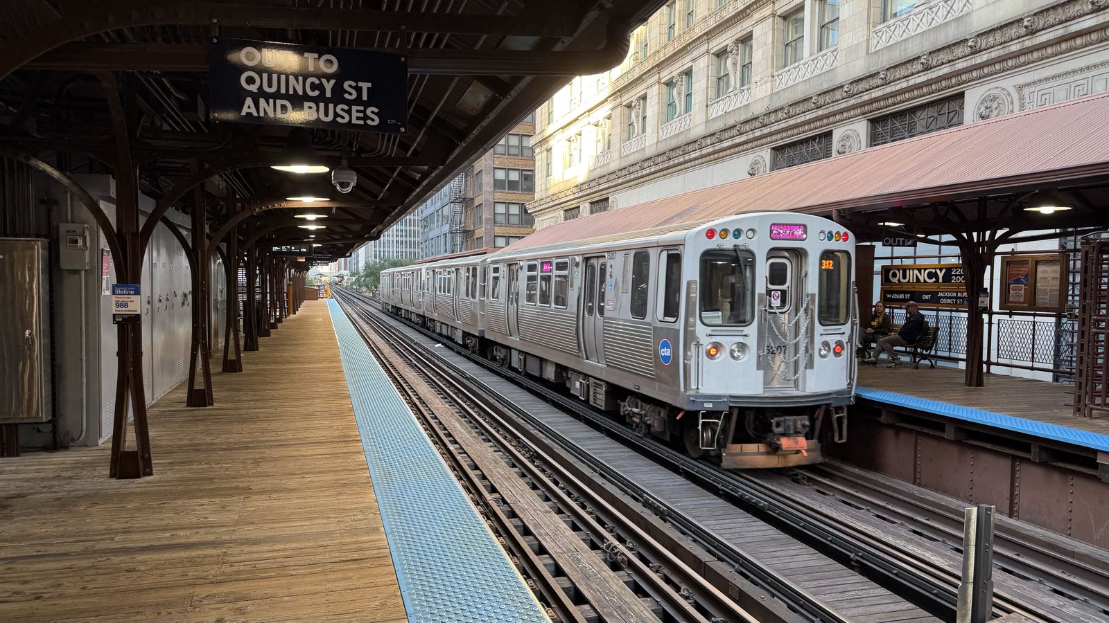 A silver subway train pulls out of an above-ground station. The station platform’s are wooden. City buildings are visible past the station platform roof. Signs indicate the name of the station is Quincy.