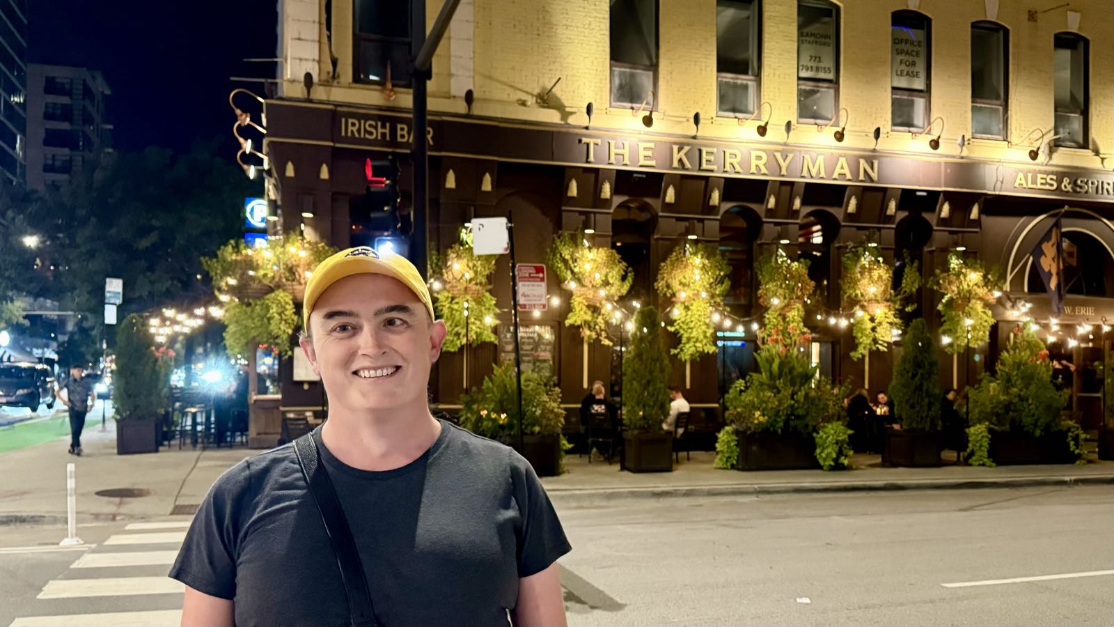 A woman in a yellow hat and gray t-shirt stands across the street from a classic-looking Irish pub called The Kerryman. The dark-paneled pub is on the ground level of a yellow-beige building, and its patio is surrounded by greenery.