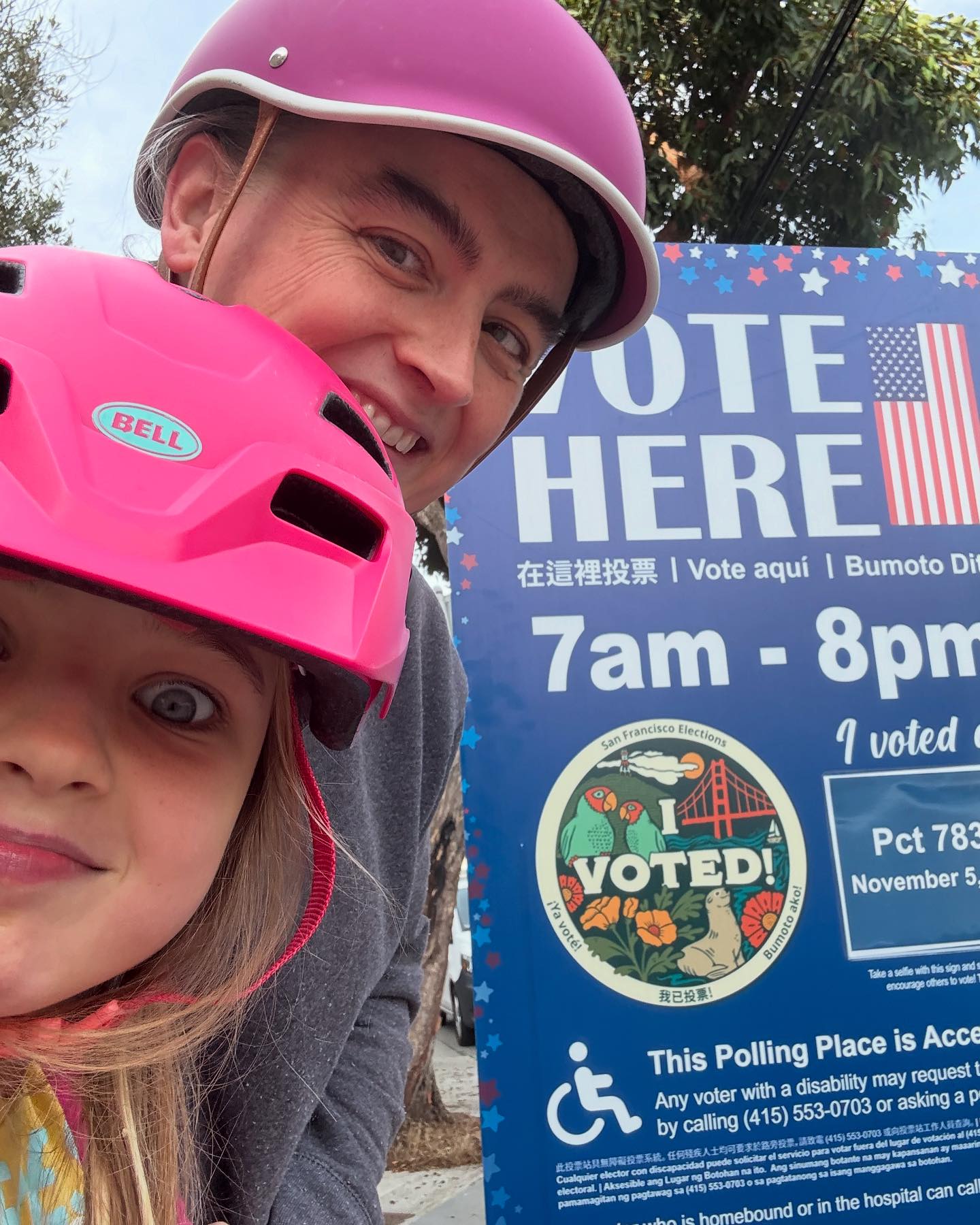 A a woman and a girl, each in bike helmets, pose before a sign designating a polling place.