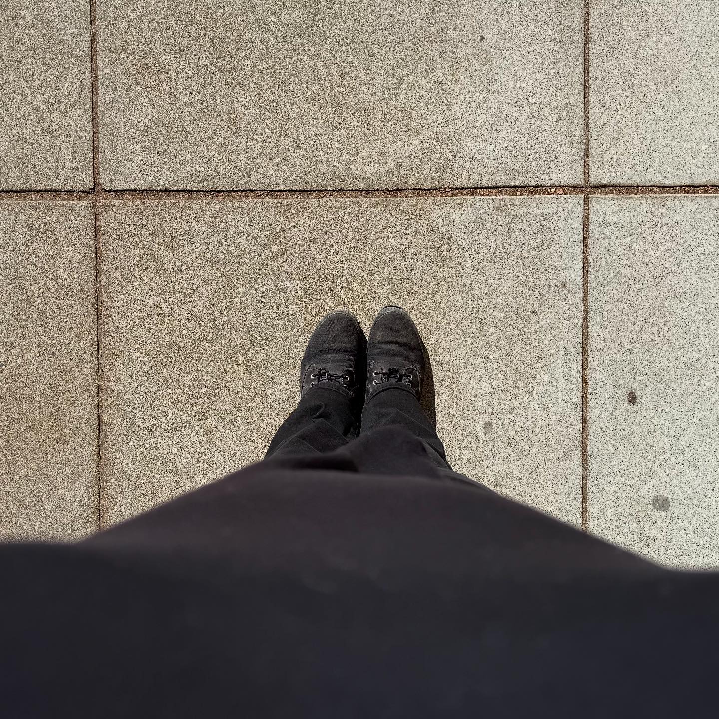A person is standing on a gray sidewalk. The photo is taken from about chest-level, pointing straight down, so we only see what the person is wearing, and not their face. They are wearing a black shirt, black pants, and black boots.