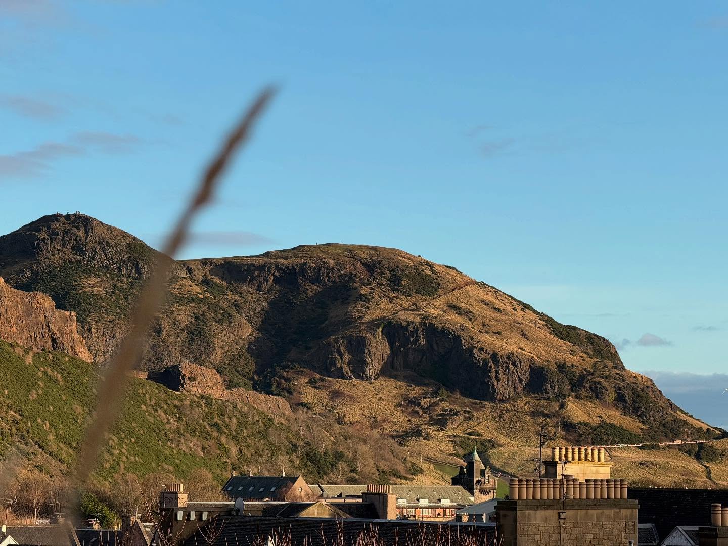 A large, brown-and-green hill looms over the tops of buildings. The sky above the hill is blue. Not much is visible of the buildings besides roofs and chimneys.