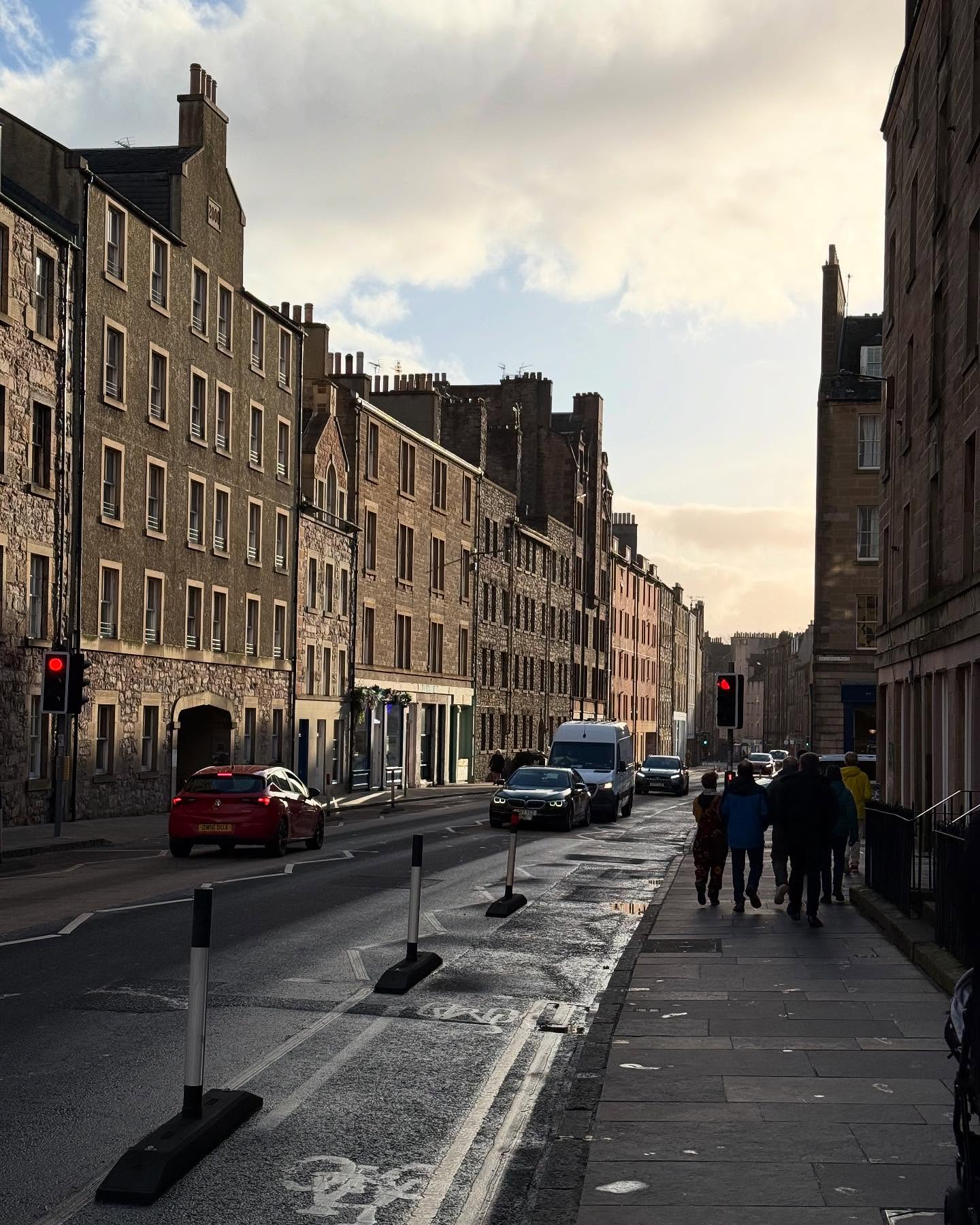 A two-way city street has a bike lane along the near sidewalk. Each side of the street is lined with gray stone buildings dating from the 18th, 19th, and 20th centuries. A traffic light is red, and cars are waiting on both sides of it. A group of people walks along the sidewalk at a distance. They’re sufficiently bundled up that it must be cold out, but there’s little else in frame to suggest the temperature.