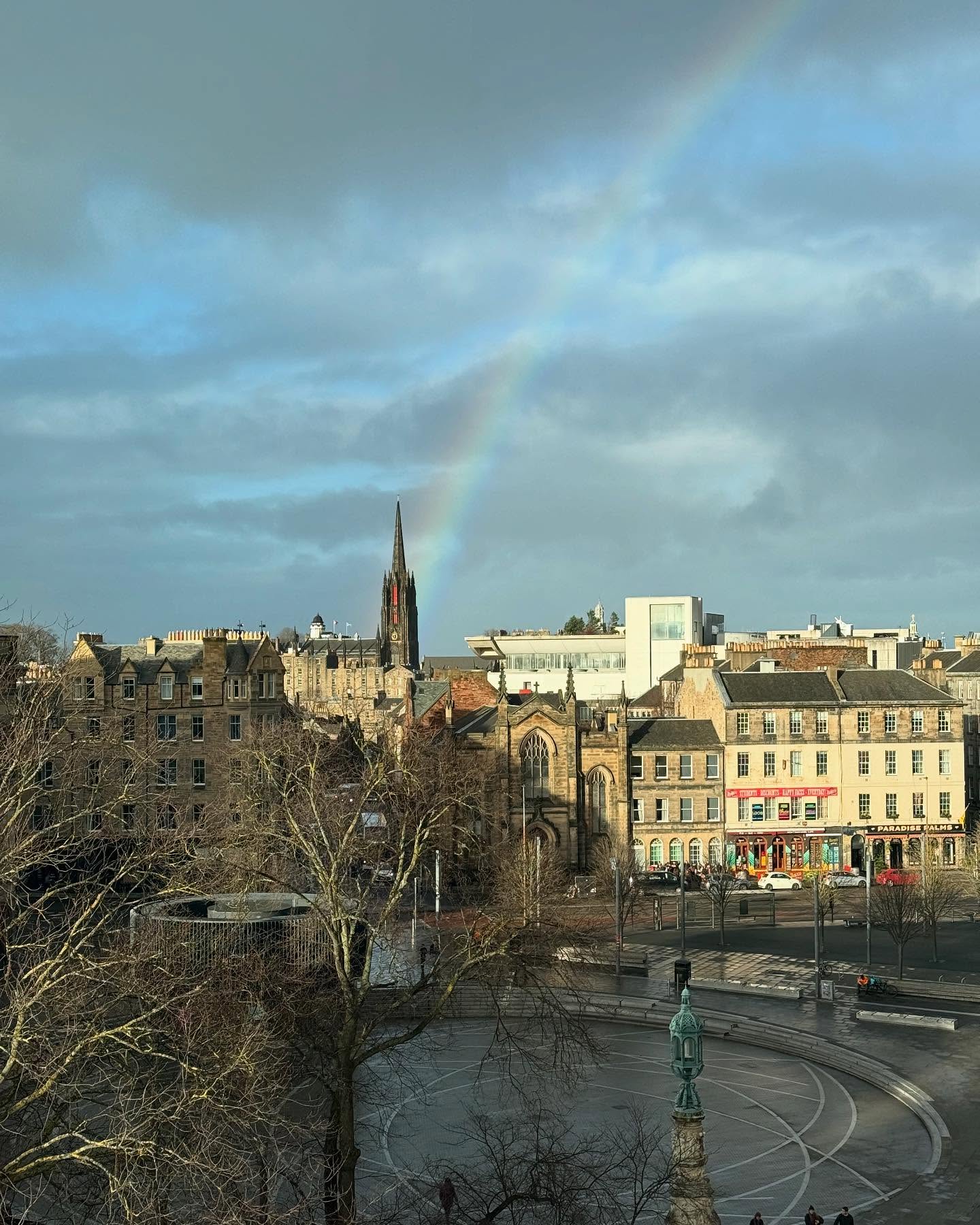 A rainbow descends from a cloudy sky, meeting the city at a dark church steeple. The portion of the city skyline that’s visible is a mixture of older and newer buildings, all various shades of gray that suggest a continuity of building material over the centuries. The church whose steeple is receiving the rainbow is at center. In between the camera and the city buildings is a large-ish cleared space, in whcih at least one statue is visible; it’s presumably a city square of some kind.