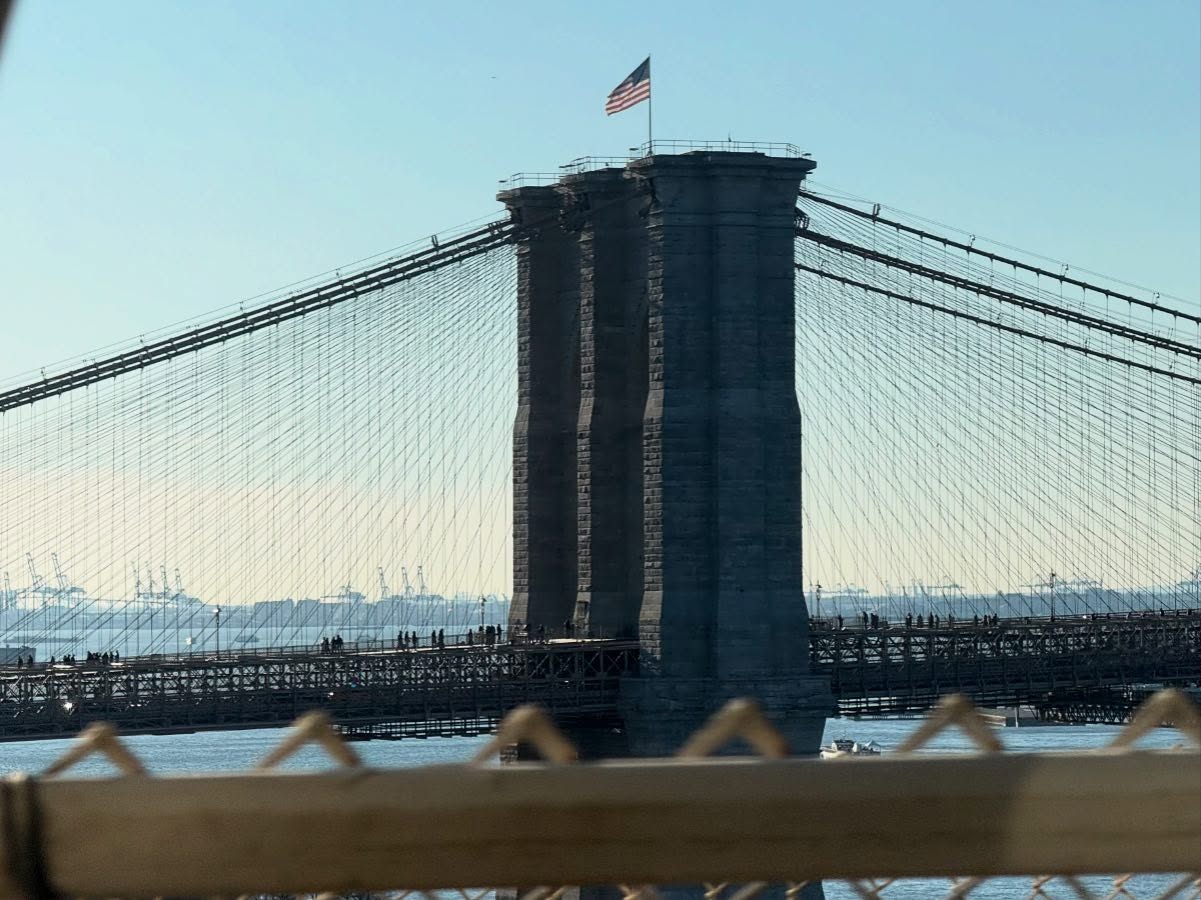 The eastern tower of the Brooklyn Bridge is in center frame, viewed from the north. Cables descend from it to either side, and an American flag flies above it.