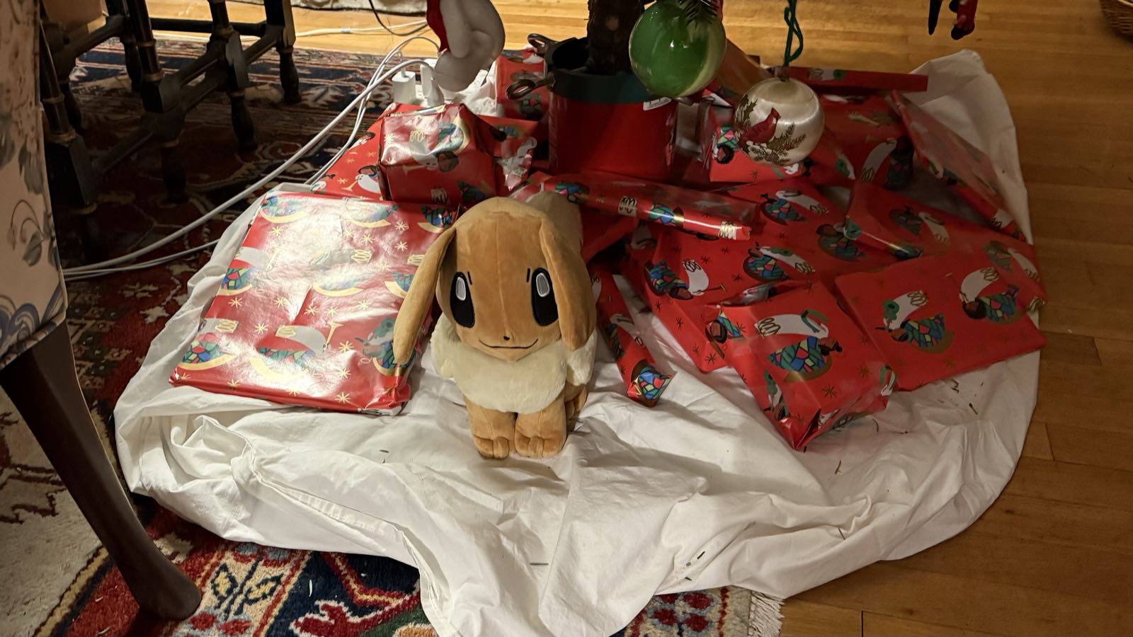 A large pile of presents underneath a Christmas tree. Most are wrapped in red wrapping paper; one, a stuffed animal (of the Pokémon Eevee) is not.