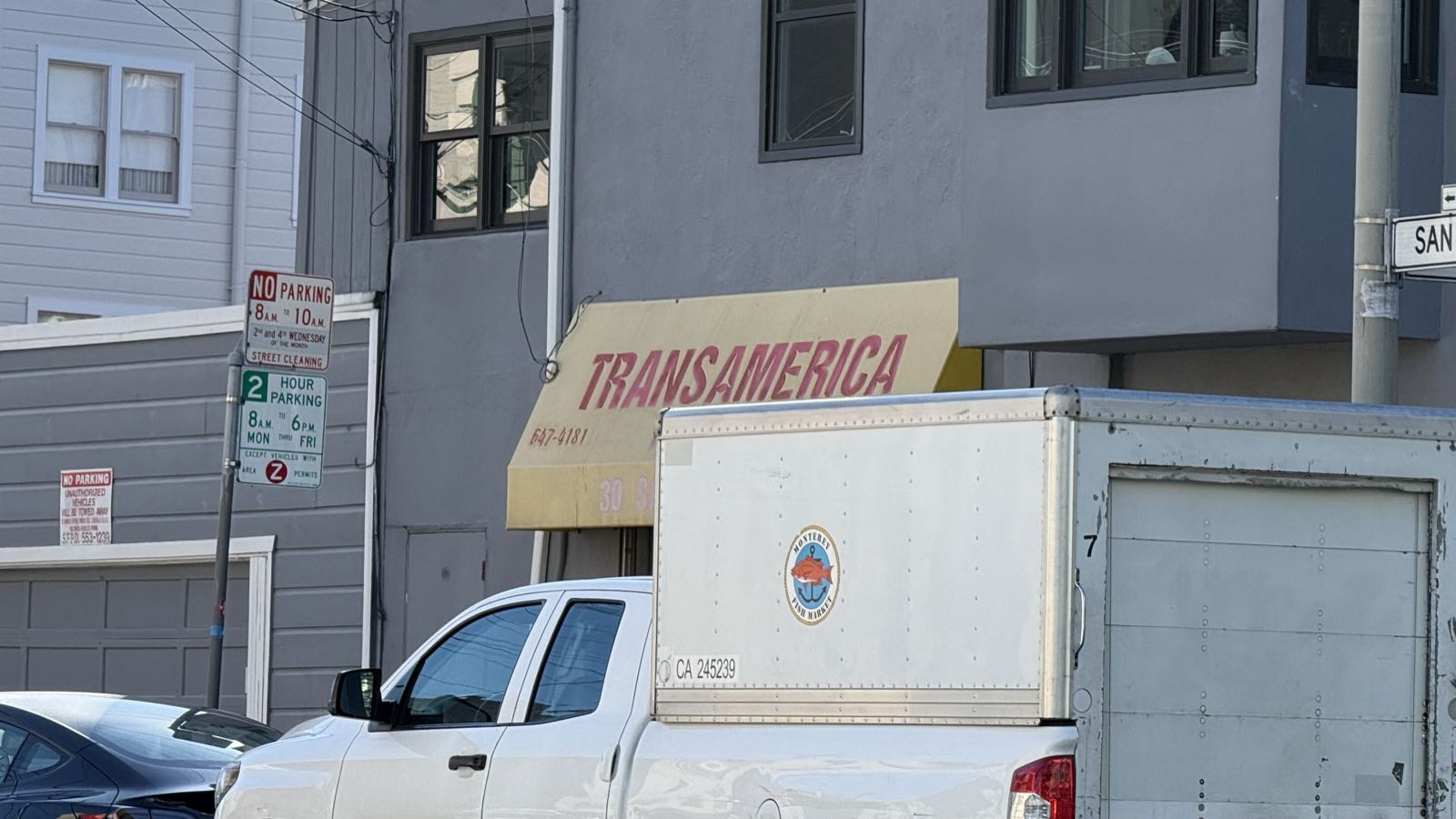 Behind a parked truck on a city street, a yellow awning is visible. The awning has “TRANSAMERICA” written on it in large, red letters.