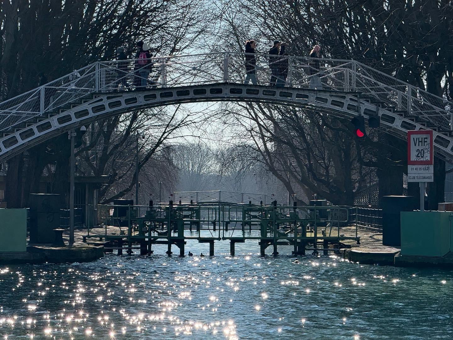 Canal Saint-Martin on a sunny day. The canal is sparkling with sunlight. A tall pedestrian bridge arches over the canal at the center of the frame. Below and beyond the bridge, some of the canal’s locks are visible.