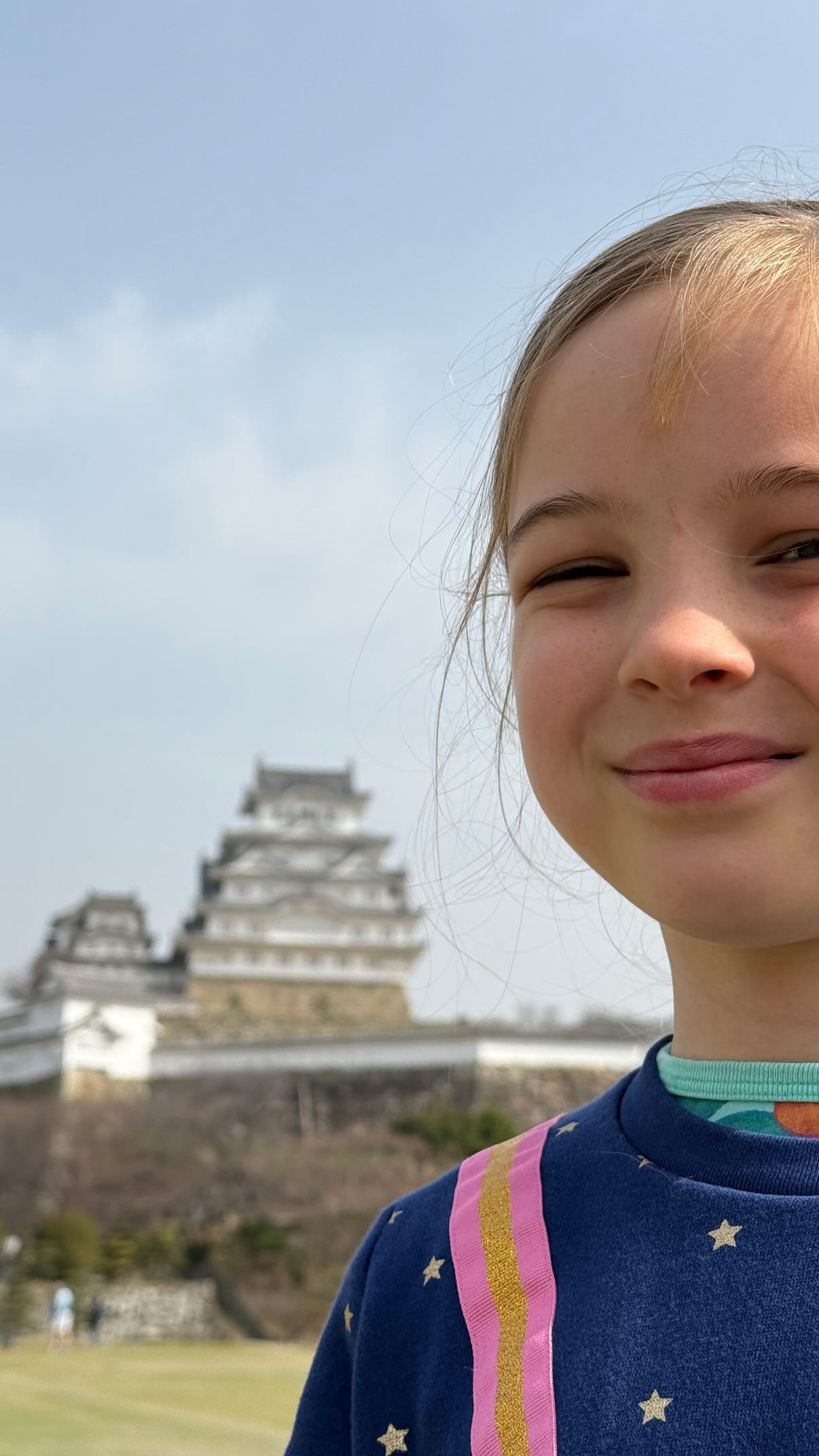 A child smiles at the camera. Behind the child, in the distance, is a white Japanese castle.