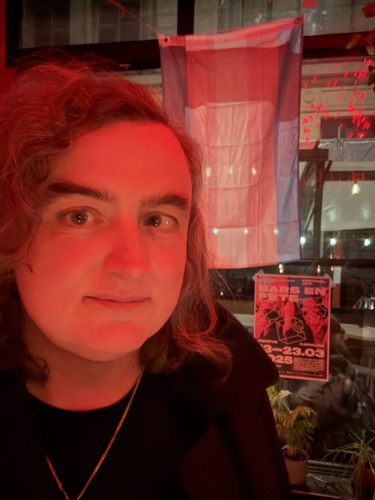 A woman stands in front of a window, in which hangs the transgender pride flag. The woman is in dark clothes, and has some kind of silver necklace on. Her shoulder-length hair is curly.