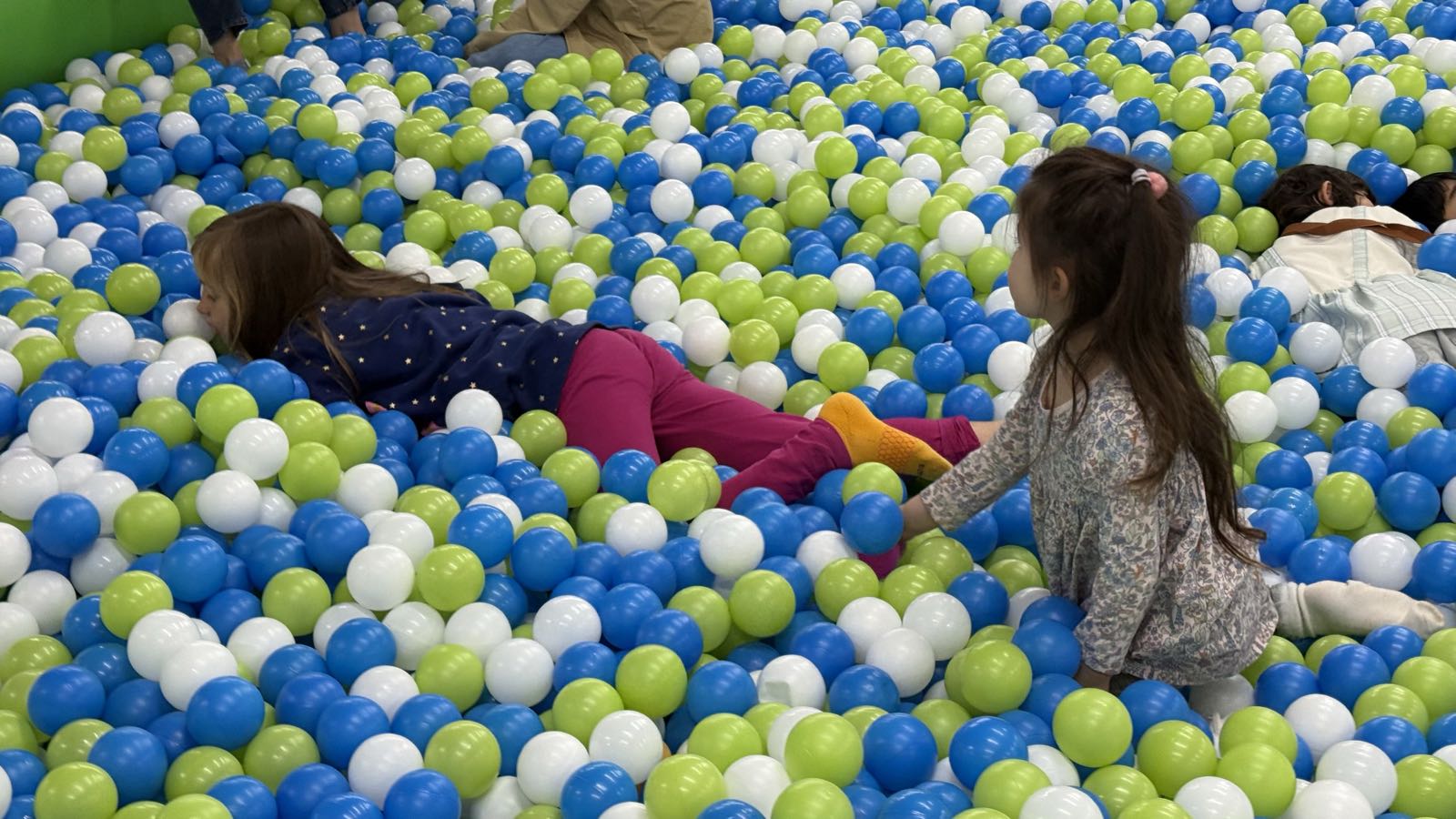 Two children play in a pool full of white, blue, and green balls. One is lying face-down, while the other is kneeling.