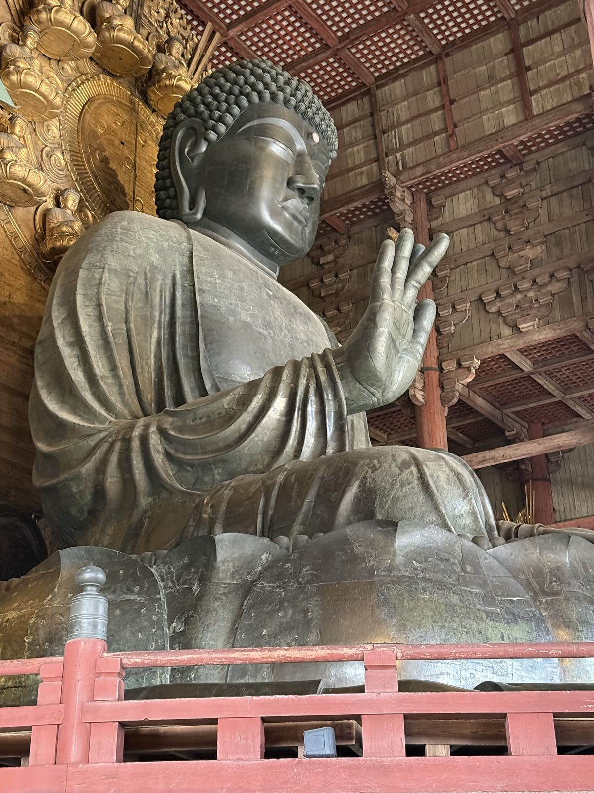 A very large bronze Buddha holds one hand up. There is a golden throne visible behind the Buddha. The room the Buddha is sitting in is elaborately made of wood.
