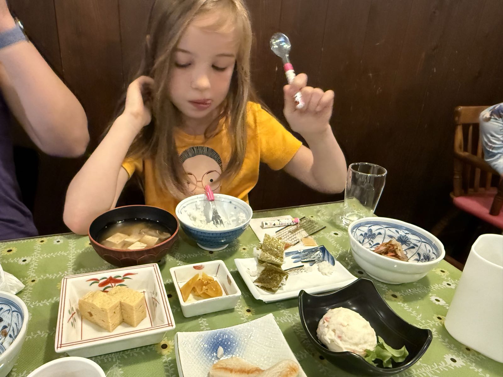 A child holds a spoon while looking down at many small plates and bowls of food in front of them.