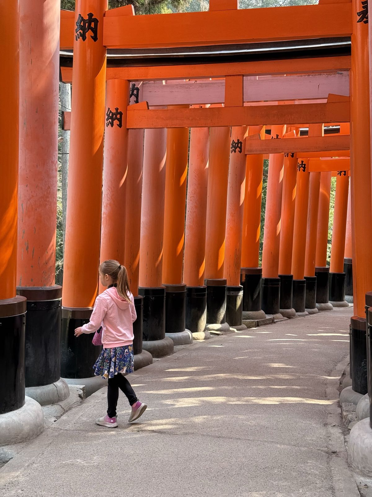 A child in a pink sweatshirt with a long ponytail walks alone in a tunnel made of many vermilion torii gates.