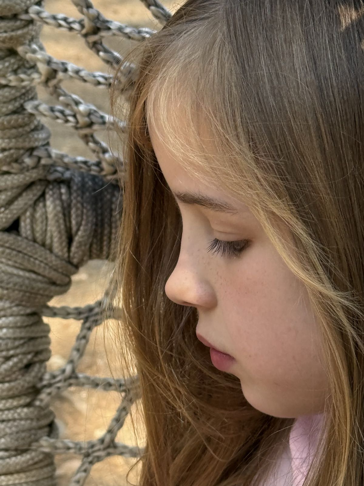 A side profile close-up of a child sitting in some kind of rope or wicker chair. The child has dirty blond hair and is sporting a neutral expression on their face.
