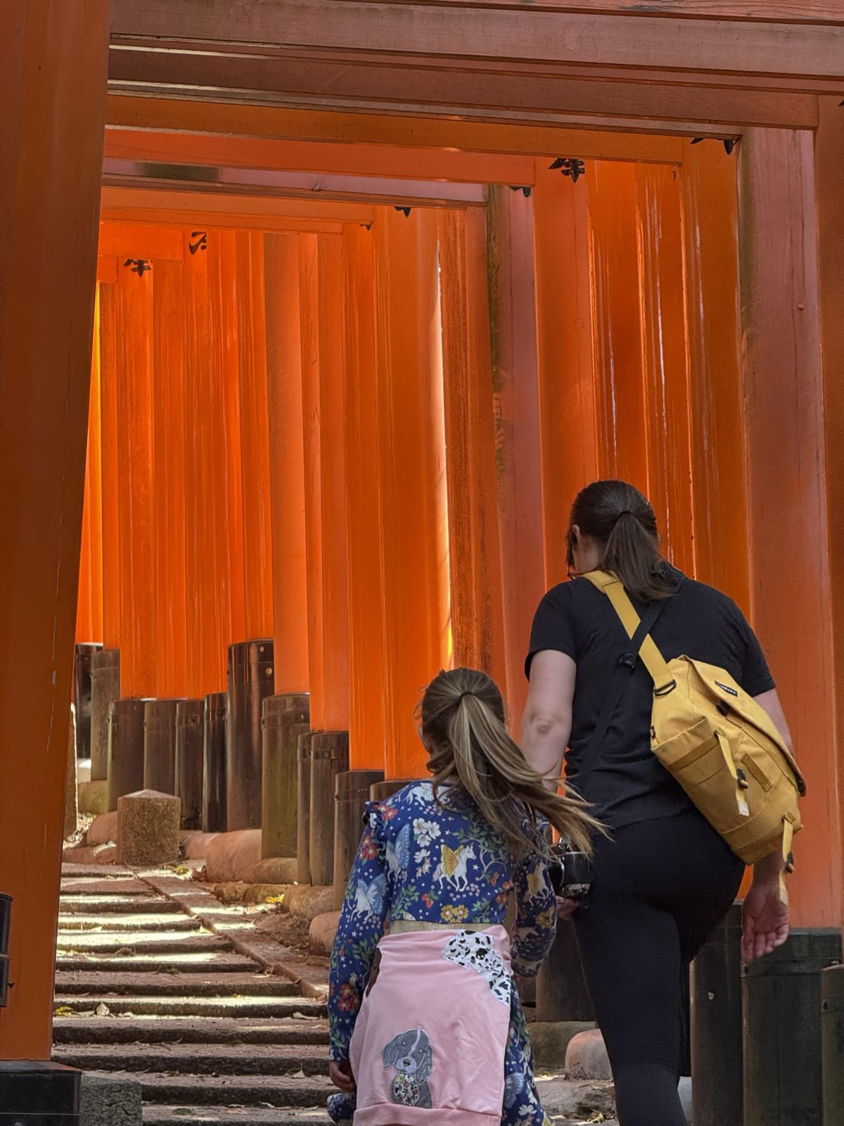 A child and a woman climb stone steps within a tunnel of vermilion torii gates. The gates are lit up by the sun in a very vibrant manner.