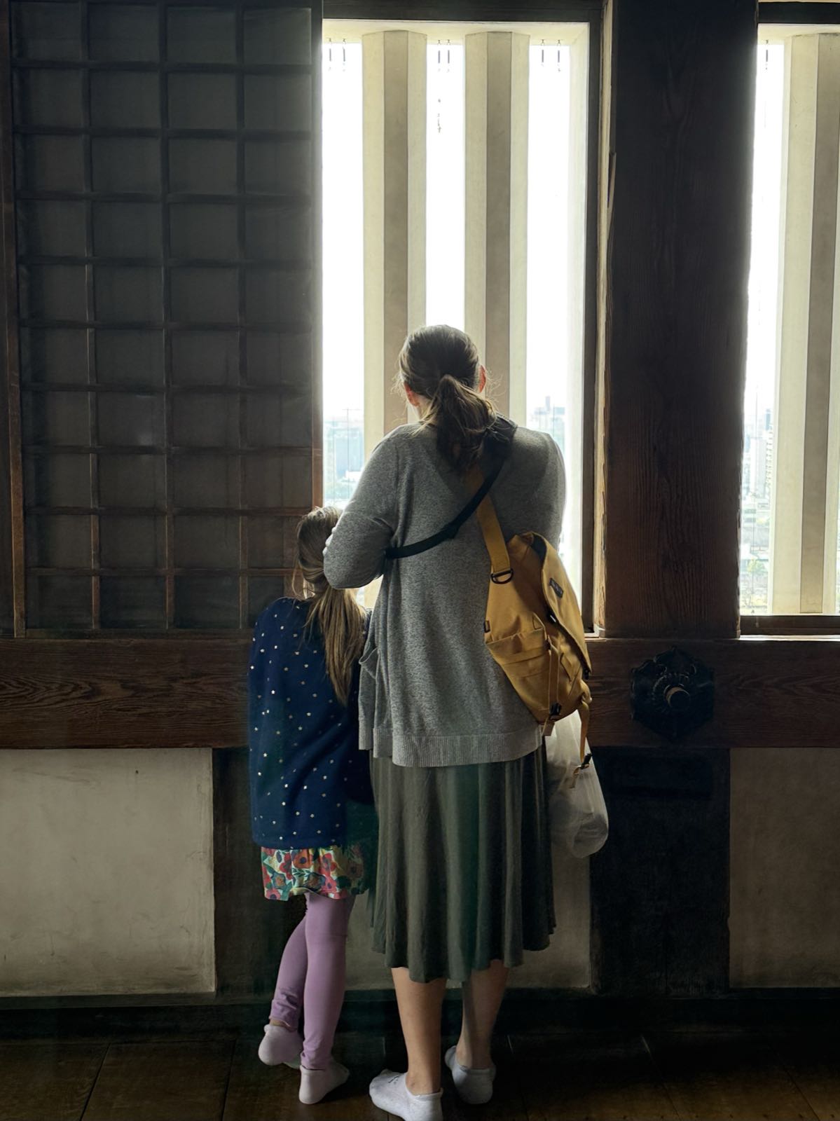 A child and a woman are inside a room, looking out a brightly-lit window. The window has narrow vertical slats. The room is of dark wood and a light colored plaster.