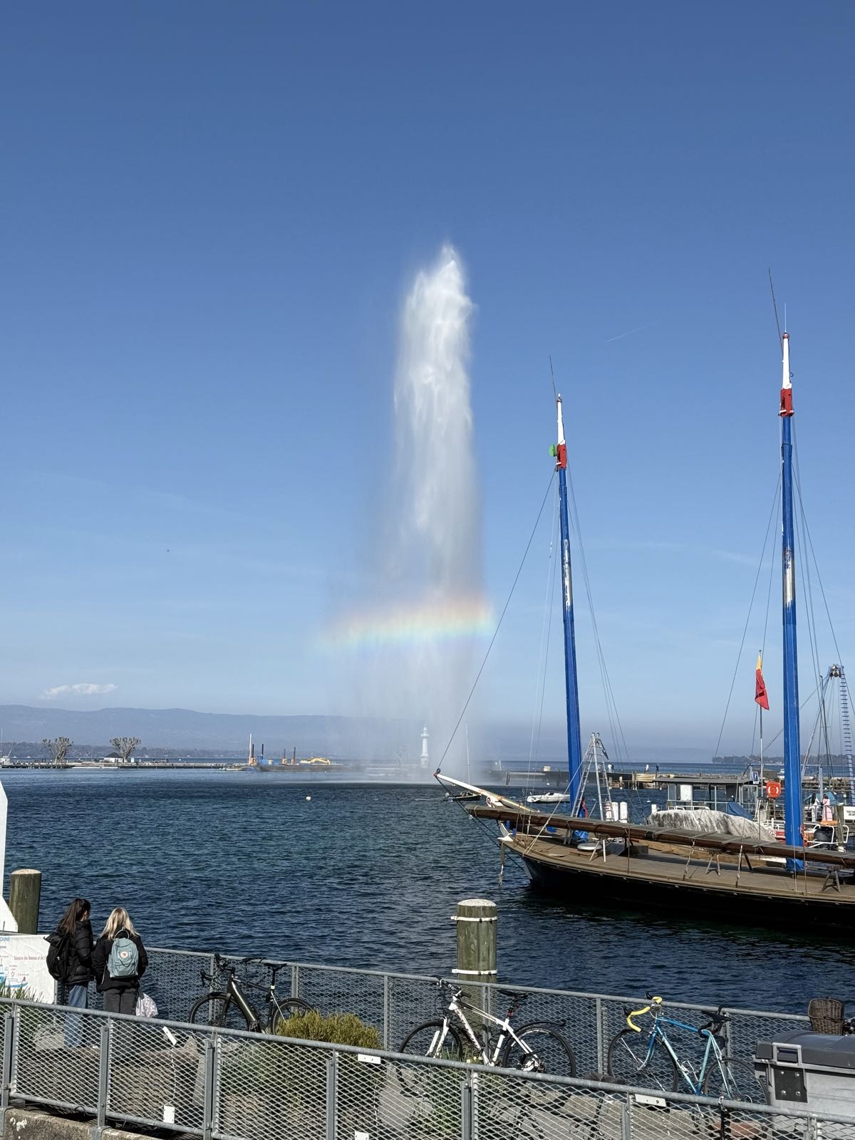 A tall plume of water is being ejected from a water fountain in the middle of a large body of water on a sunny day. There is a rainbow visible in the stream of water. In the foreground, some boats and a pier are visible.
